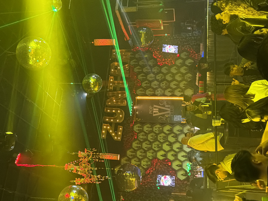 Inside a nightclub, "Zuma" is displayed in large, illuminated letters alongside "Pop Az Club." Disco balls hang from the ceiling, reflecting bright yellow spotlights and green laser beams. The walls feature a honeycomb pattern adorned with miniature bottle-shaped decorations and splashes of red floral elements. Patrons are seated at tables, partially visible amidst the vibrant lighting and energetic atmosphere. A decorative chandelier-like fixture hangs above the disco balls.