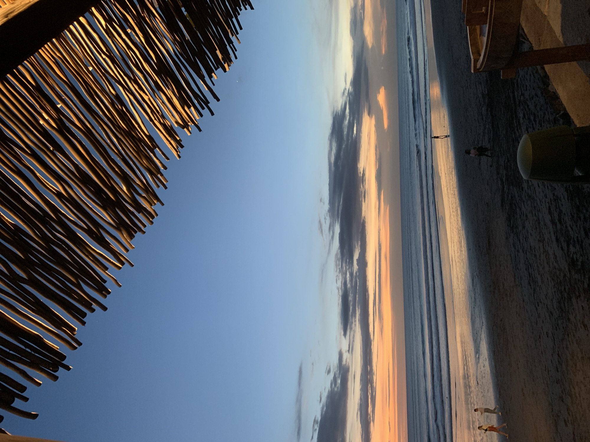 The image captures a beach scene in North Kuta, Indonesia, during what appears to be a sunset. A thatched roof or awning is prominently featured in the upper left, partially obscuring the sky. The sky exhibits a gradient from blue to shades of orange and gray, with clouds visible. The beach itself is visible to the right, with the ocean extending into the distance. A few individuals can be seen walking along the shoreline. The overall lighting indicates that it is either dusk or dawn, and the orientation of the image is rotated, with the ground appearing at the right side.