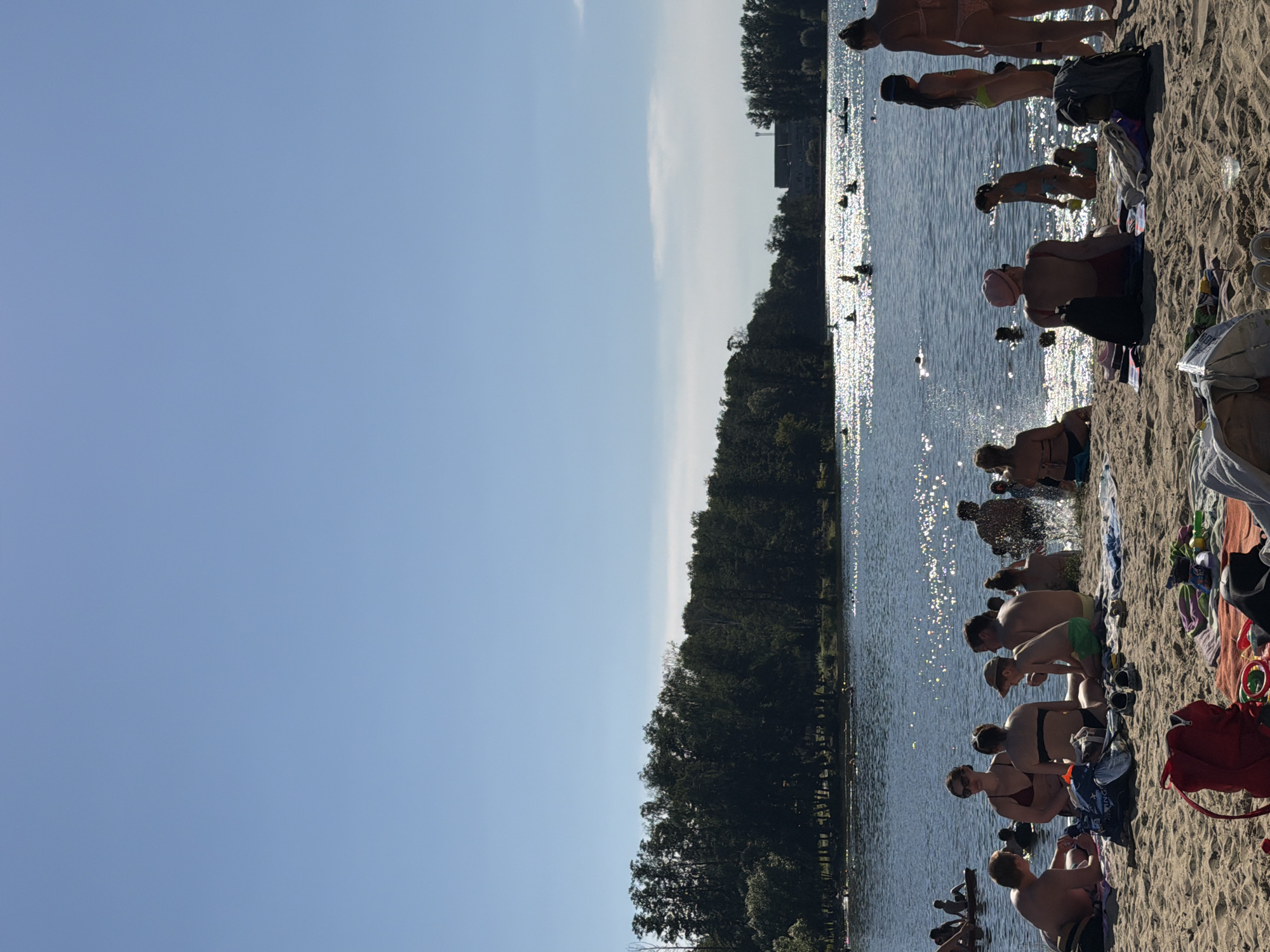 The image depicts a crowded beach scene on a sunny day, likely at a lake or reservoir within Malakhovka, Russia. The foreground is occupied by a sandy beach where several groups of people are relaxing on towels and blankets. There are various objects scattered on the sand, including bags, shoes, and toys.
In the mid-ground, a body of water is visible, with numerous people swimming, wading, and participating in water activities. The water is sparkling under the bright sun, and some individuals appear to be on inflatable devices or small boats. The shoreline is lined with a dense forest of green trees.
In the background, across the water, there are buildings visible, suggesting the presence of a developed area near the recreational spot. The sky is a clear, bright blue with a few faint clouds. The overall atmosphere is one of leisure and summer recreation.