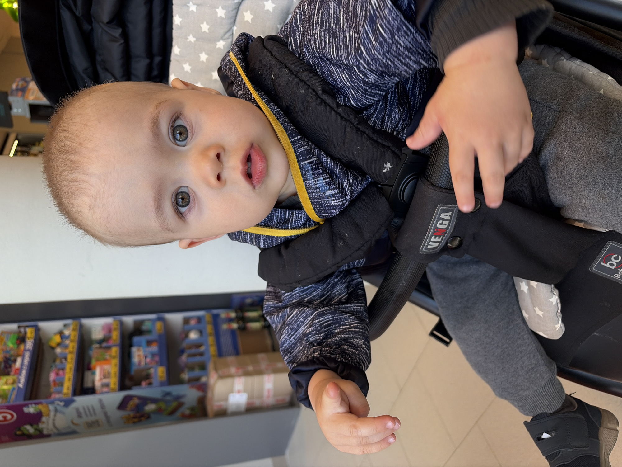 A baby with fair skin, grey eyes, and light blonde hair is sitting in a VEGA stroller. The baby wears a navy blue patterned jacket with a yellow trim and grey sweatpants with black sneakers. The stroller has a black harness with a buckle. A shelf with toys in boxes is visible in the background. The location is likely Chaikovsky, Russia.