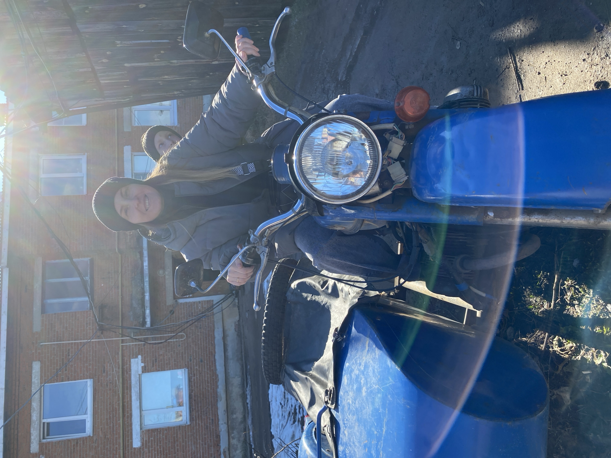 Two people are on a blue motorcycle with a sidecar in Noviy, Russia. The motorcycle is facing the viewer with the headlight prominently visible. A woman with dark hair and a black beanie is sitting on the driver’s seat, smiling. Behind her, a second person with a dark beanie is partially visible. The backdrop features a brick building with several windows and a wooden structure. The ground appears to be a mix of dirt and foliage, suggesting an outdoor setting. The lighting is bright, possibly indicating a sunny day.