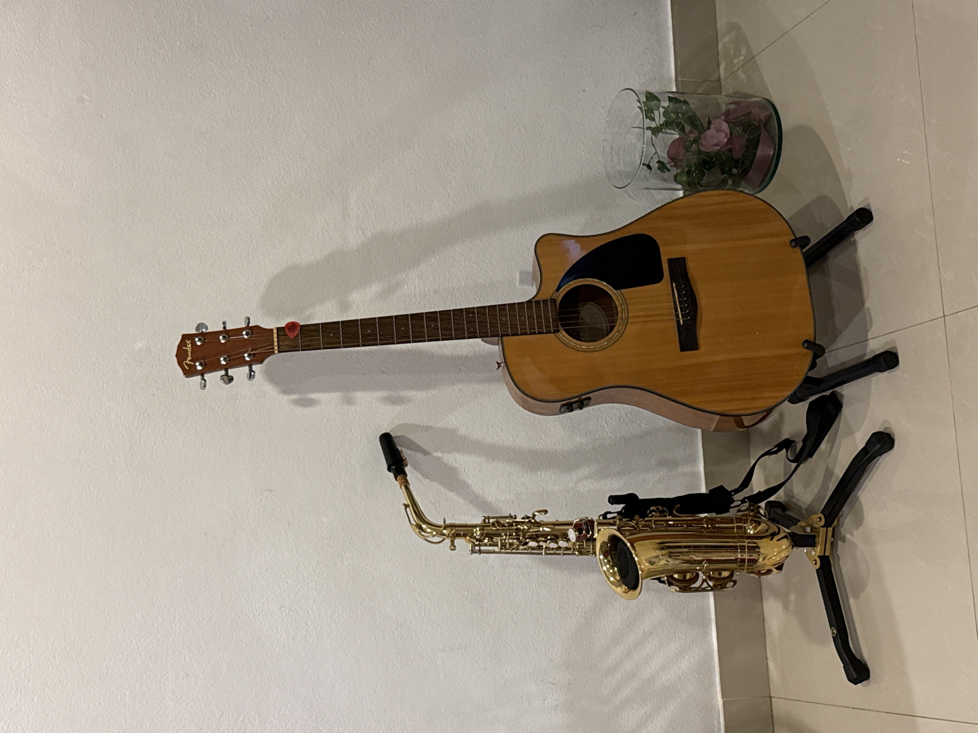 The image captures an indoor scene in Baan Chaweng Noi, Thailand, featuring musical instruments against a plain white wall and light-colored tiled floor.
The primary subjects are a light-brown acoustic guitar and a gold-colored alto saxophone. The acoustic guitar stands upright on a black, multi-legged guitar stand. The brand "Fender" is visible on the guitar's headstock, and a small red guitar pick is tucked into the strings near the tuning pegs. The guitar has a cutaway body and a dark pickguard.
Adjacent to the guitar, a shiny gold alto saxophone rests on a black saxophone stand. The saxophone has a black mouthpiece and a strap attached to its body.
In the background, positioned near the top of the guitar, is a clear glass vase or bowl containing artificial pink roses and green foliage. Shadows from the instruments and the vase are cast distinctly on the white wall, indicating an indoor light source. The floor consists of large, light-colored square tiles. The overall scene suggests a static display or storage arrangement for the musical instruments. There are no people visible, and no specific activity is taking place.