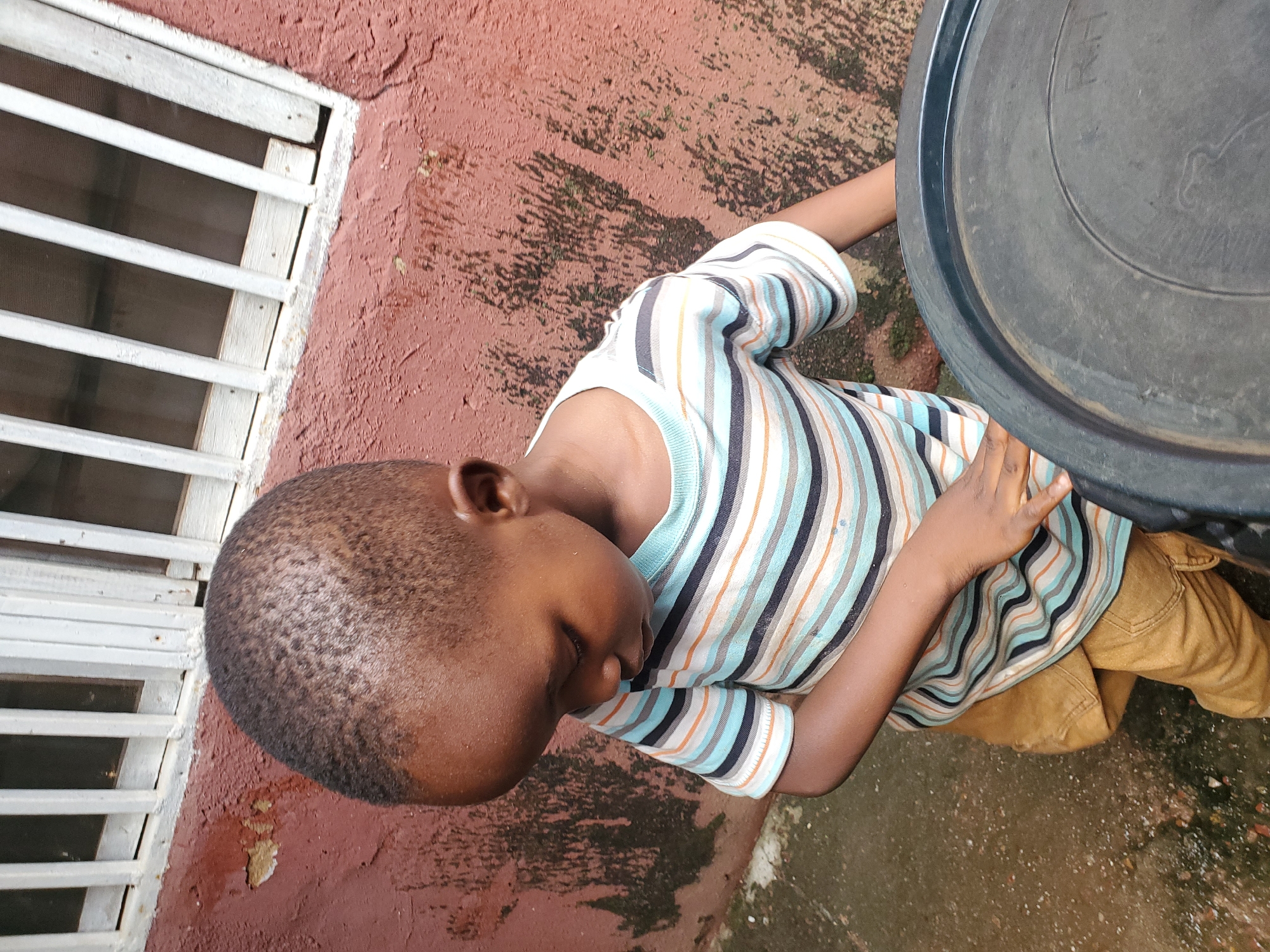 The image shows a young boy wearing a striped t-shirt and brown pants. He is lying down on a reddish-brown textured surface, possibly a wall or ground. To his left, there is a white slatted window or barrier. To his right, a large, dark, circular object, possibly a bin or container, is visible. The scene appears to be outdoors, with some dirt and debris on the ground. The lighting suggests it could be daytime. The boy's eyes are closed, and his head is tilted downwards, giving the impression that he is resting or possibly asleep. The overall mood is one of quiet repose. There is no visible text in the image. The location context provided is Jos, Nigeria.