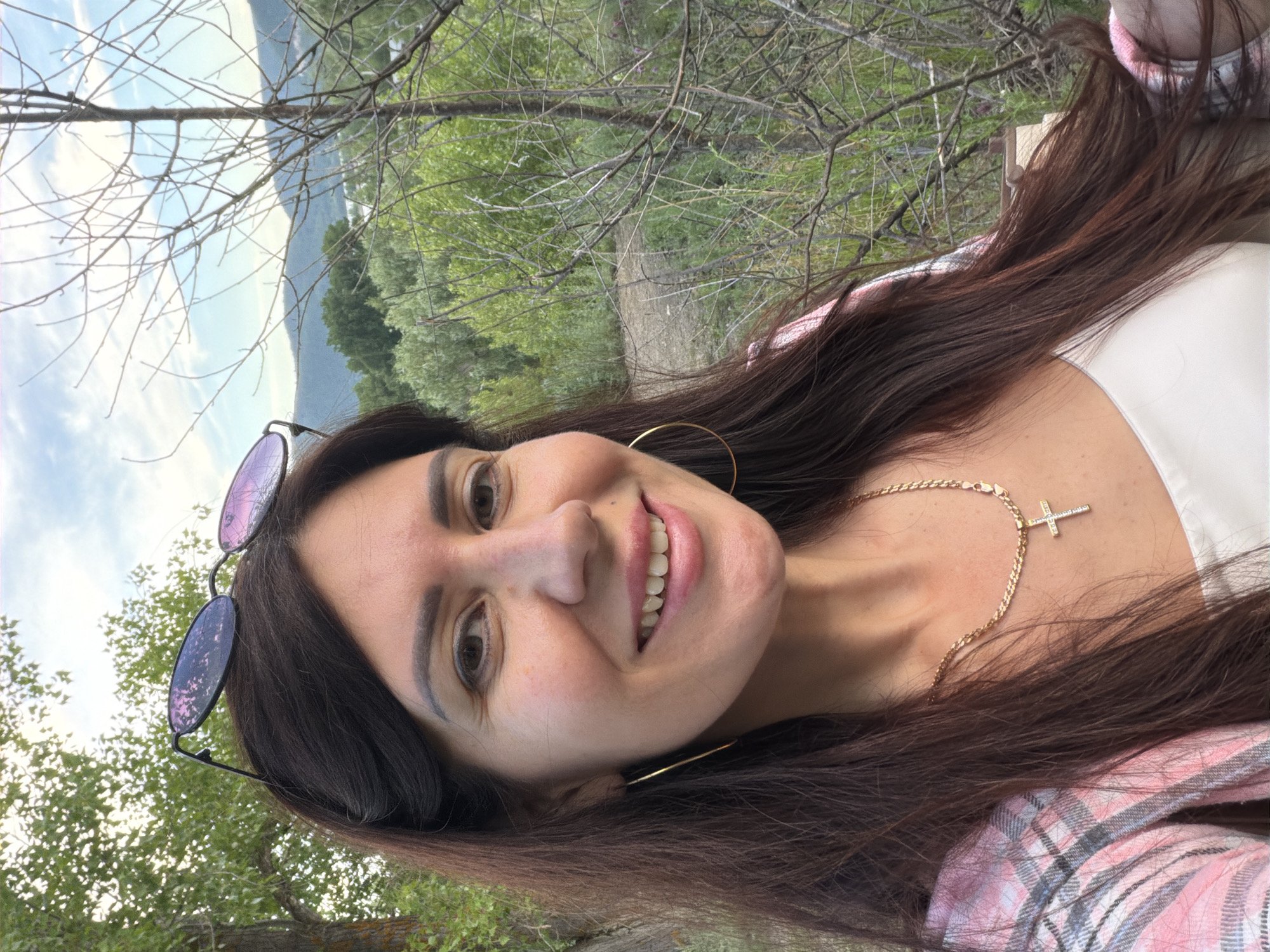 In a close-up selfie taken outdoors, a woman with long, dark brown hair smiles at the camera. She is wearing a white top, a pink and gray plaid jacket, a gold necklace with a cross pendant, and large gold hoop earrings. Sunglasses with purple lenses are perched on top of her head. The background is a natural landscape with trees, foliage, and a visible hillside under a cloudy sky. The image appears to be taken during daylight. The location is not discernible from the visual information provided.