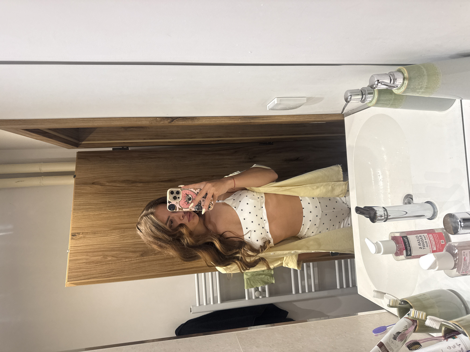A young woman with long, wavy brown hair takes a mirror selfie in a modern bathroom. She is wearing a white polka dot bralette and matching shorts, with an open light yellow button-down shirt draped over her shoulders. Her phone, with a decorative case featuring an eye and stars, is held up to capture the reflection. The bathroom features a wooden-paneled wall, a white sink with a chrome faucet, and various toiletries on the counter, including toothbrushes in a holder and pump bottles of soap and lotion. In the background, a white towel rack with a towel is visible against a light-colored wall. The scene appears to be in a residential bathroom. The image does not contain any specific visual cues that would allow for localization to Plovdiv, Bulgaria.