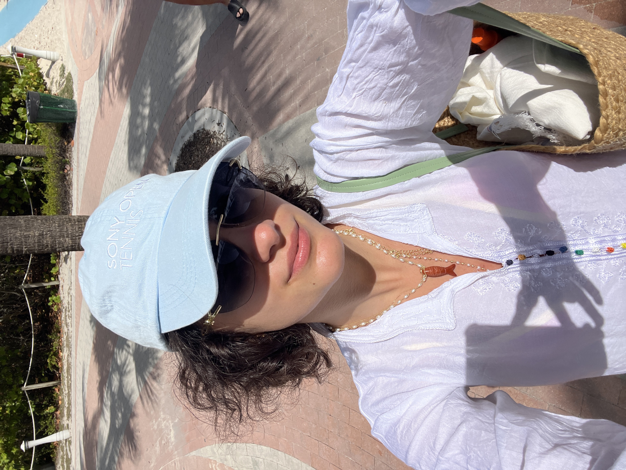 A medium-level, eye-level shot showcases a woman in Miami Beach, Florida, in the United States, dressed in a white, long-sleeved top, a light blue baseball cap with "SONY OPEN TENNIS" embroidered on it, and large sunglasses. She also sports a gold necklace with a pearl strand and a fish pendant. A straw tote bag with white fabric inside is slung over her shoulder. The backdrop consists of brick ground, a palm tree, a trash can, and a glimpse of foliage on a sunny day.