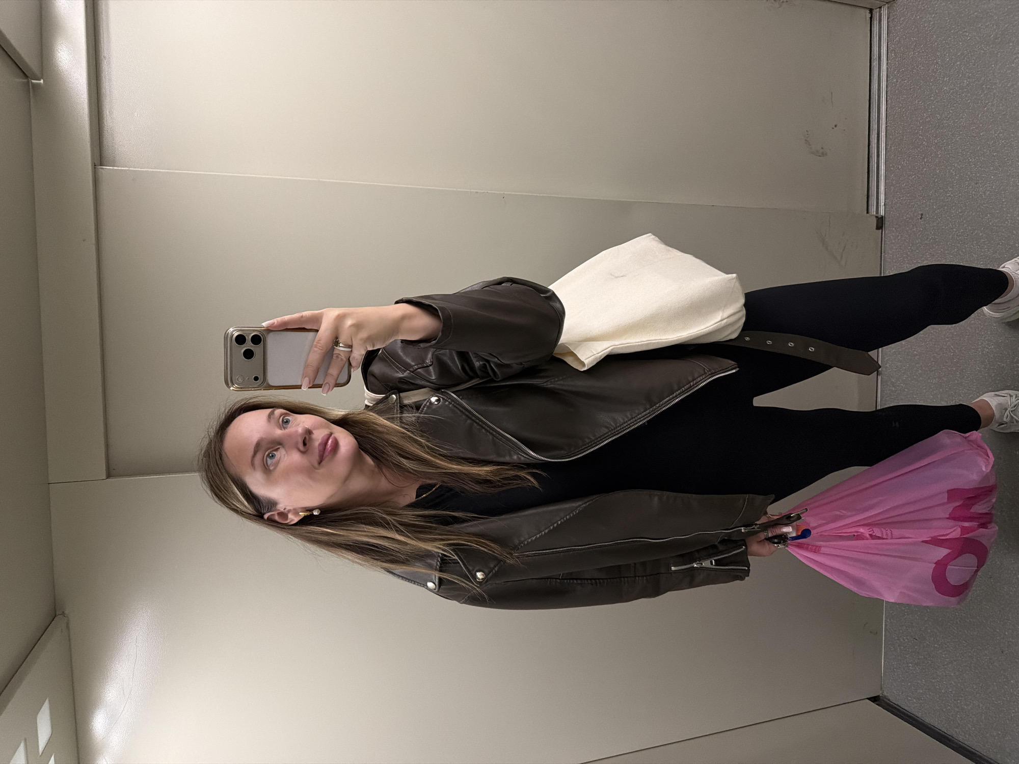 A woman is taking a selfie in what appears to be an elevator. She is wearing a brown leather jacket, a black top, and black leggings. She's holding a phone in her left hand and a pink plastic shopping bag in her right hand. A white tote bag is slung over her shoulder. The elevator interior is plain with a metallic finish on the doors and light gray walls. The location could potentially be in Chaikovsky, Russia, although there is no definitive visual confirmation in the image.