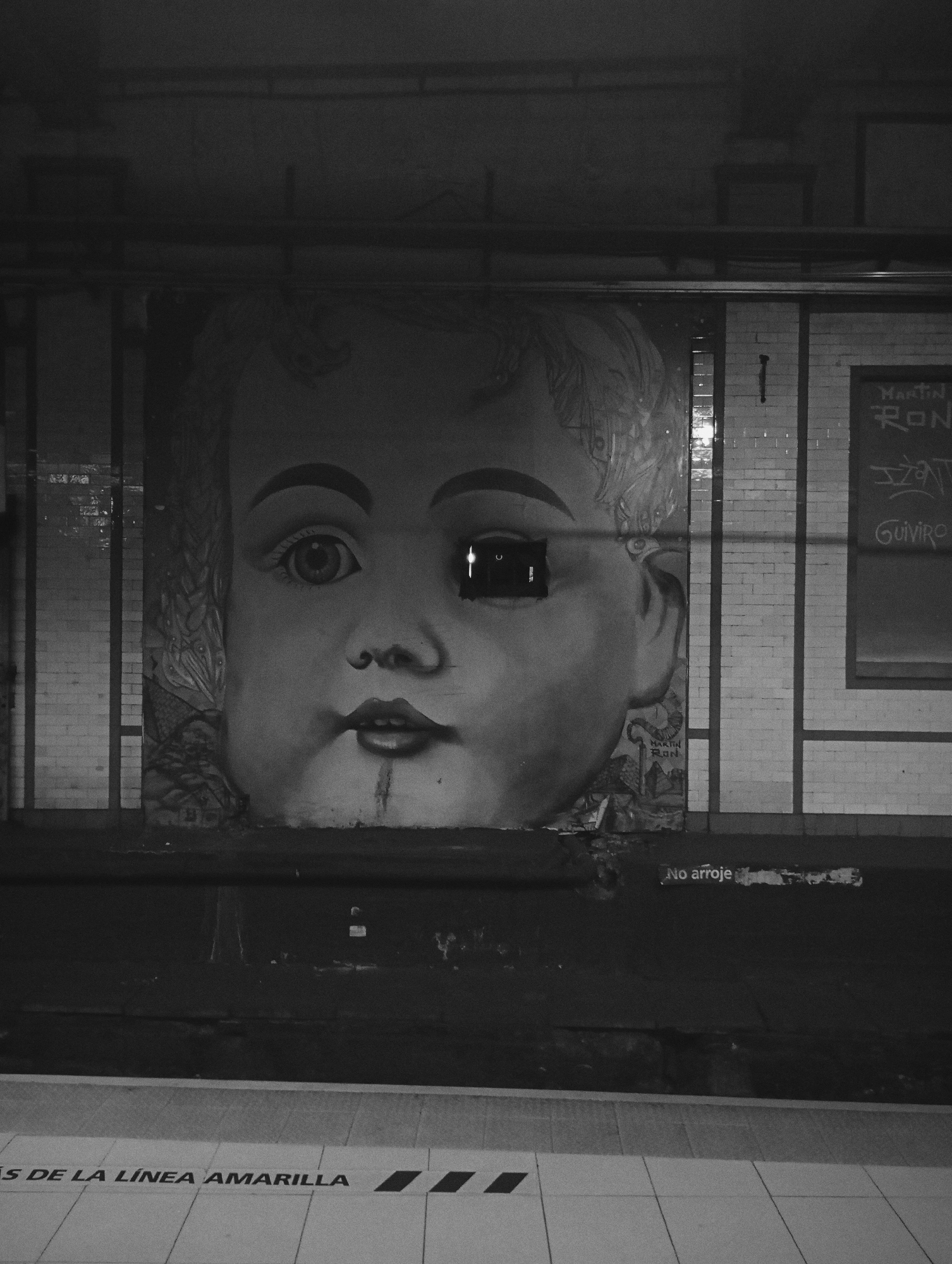The image presents a dimly lit, monochrome photograph of an underground subway station platform at Estación Carlos Gardel of Line B (Línea B) in the Buenos Aires Subte, Buenos Aires, Argentina. Dominating the wall across the tracks is a large-scale mural of a baby's face, rendered in light gray tones with prominent dark outlines. The artist's signature, "MARTIN RON," is visible near the bottom right of the baby's face. The baby's left eye (viewer's right) is recessed by a dark, rectangular void with a distinct vertical light reflection within it. A dark, vertical drip mark is present directly below the baby's slightly parted lips.
The wall behind the mural appears to be constructed of large, light-colored tiles. To the right of the mural, the wall transitions to smaller, rectangular white subway tiles, exhibiting visible staining and discoloration. This section features faded graffiti, including "MARTIN," "RON," and "GUIVIRO," enclosed within a dark rectangular outline, possibly an old advertisement space. Below this, at track level, the partial Spanish text "No arroje" (Do not litter) is discernable.
The foreground depicts the station platform, tiled with light-colored squares. Dark, rectangular safety markers are positioned along the platform edge. Across these tiles, the Spanish text "MÁS DE LA LÍNEA AMARILLA" (More of the Yellow Line) is clearly visible. The area immediately below the wall mural and platform edge, representing the track bed, is dark and undefined. Overhead, several dark pipes or conduits run horizontally across the upper portion of the scene. No individuals are present in the frame.