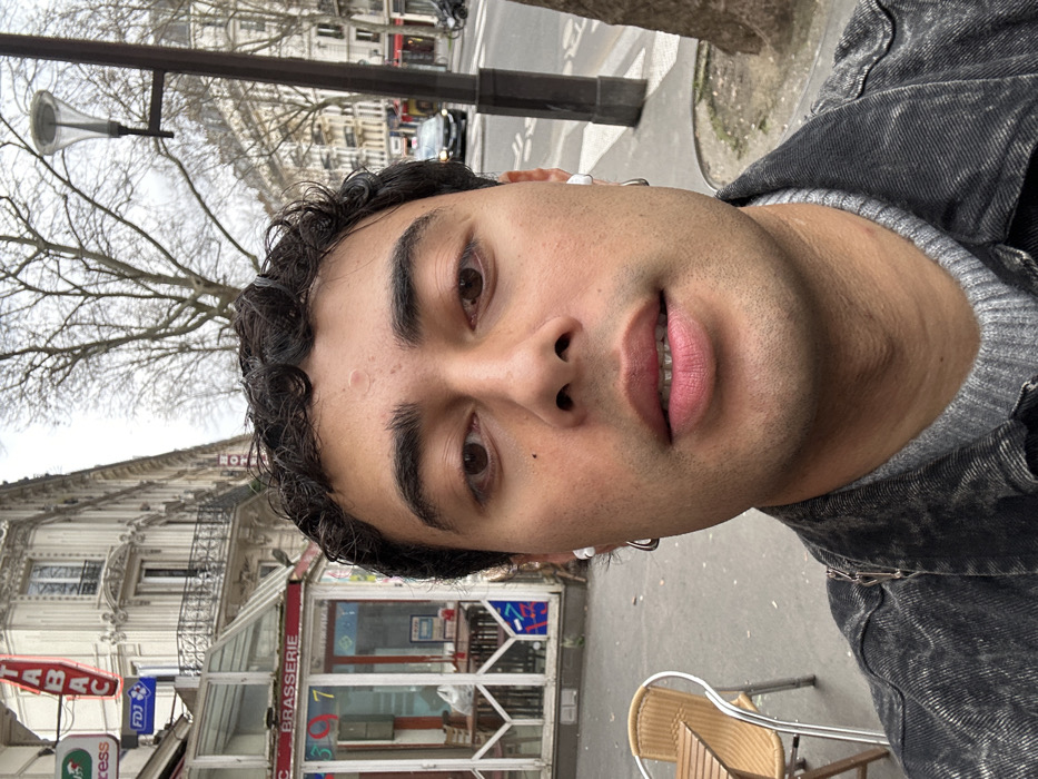 The image displays a close-up selfie of a young adult male on a street in Paris, France. The subject, positioned centrally, has dark curly hair, brown eyes, light skin, and minimal facial hair, with a small blemish visible on his forehead. He wears white wireless earbuds in both ears, a silver hoop earring in his left ear, a grey ribbed sweater, and a dark grey or faded black denim jacket. A silver chain is visible on his left shoulder. His mouth is slightly open, and he looks directly at the camera.
The background reveals a Parisian urban environment under an overcast sky. To the subject's left, a "BRASSERIE" is visible with a glass-enclosed outdoor seating area and a solitary empty cafe table with two wicker chairs on the pavement. Above the brasserie, a vertical red neon sign spells "TABAC," and a blue "FDJ" sign is also present. Behind these establishments is a multi-story Haussmannian building characterized by ornate balconies. To the subject's right, a street recedes into the distance, lined with bare trees, classic street lamps, and additional Haussmannian buildings. A dark-colored vehicle is partially visible further down the street, and the trunk of a tree is present in the far right foreground.