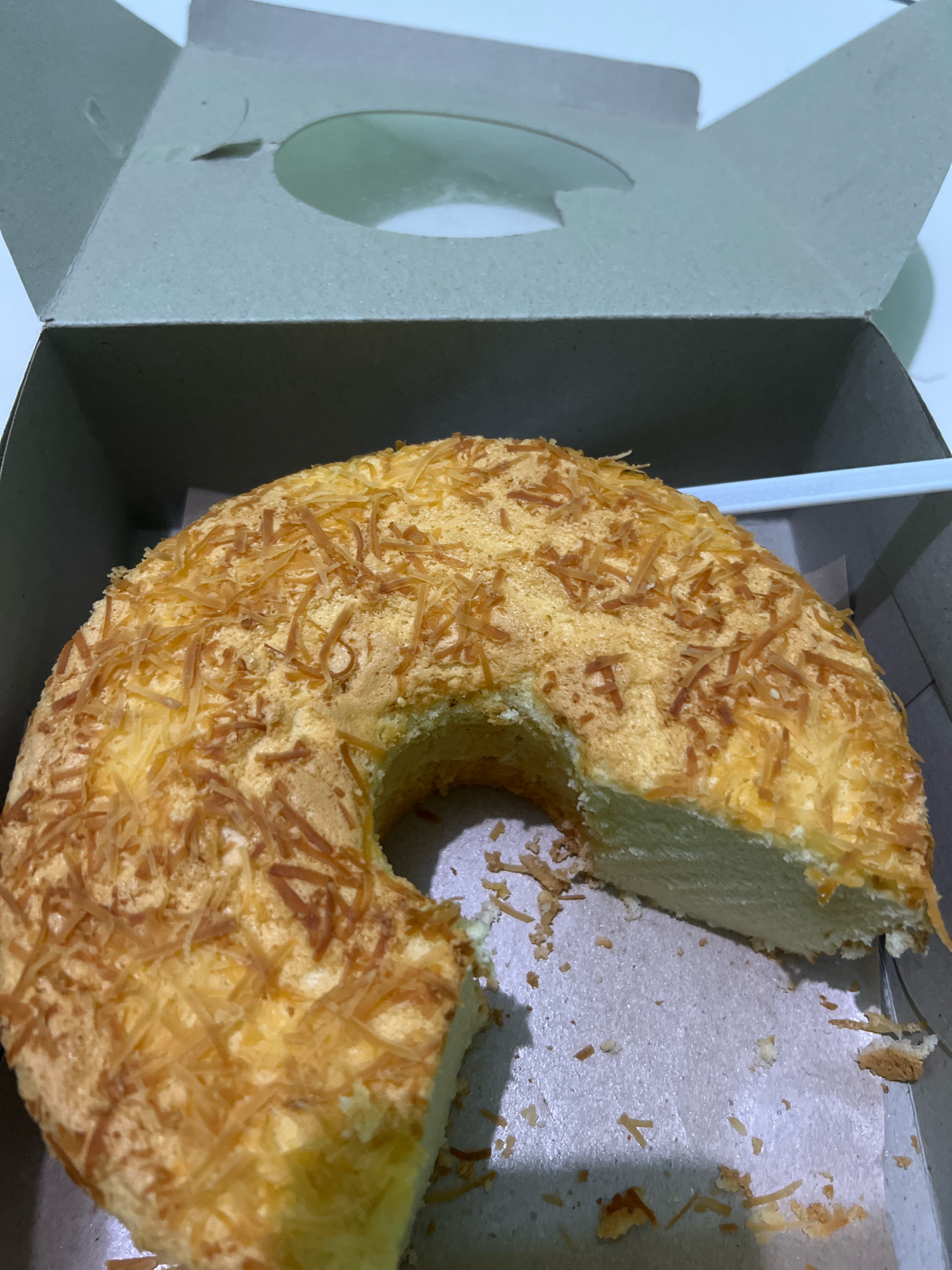 A bundt cake topped with shredded cheese sits in a gray cardboard box. The cake has a slice removed, revealing its light yellow interior. The box is open, with its lid propped up behind the cake. The scene appears to be indoors, possibly in a home or bakery in Tlogoadi, Indonesia. There are no people visible in the image.