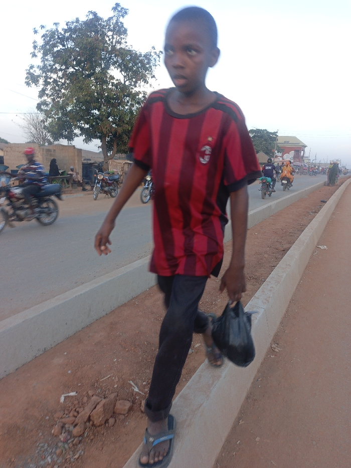 A boy wearing a red and black striped shirt, black pants, and sandals walks along a concrete curb, carrying a black plastic bag. In the background, a busy street features motorcycles and people. A tree and buildings are visible on the other side of the road. The setting appears to be a populated area, possibly a market or town, in daylight.