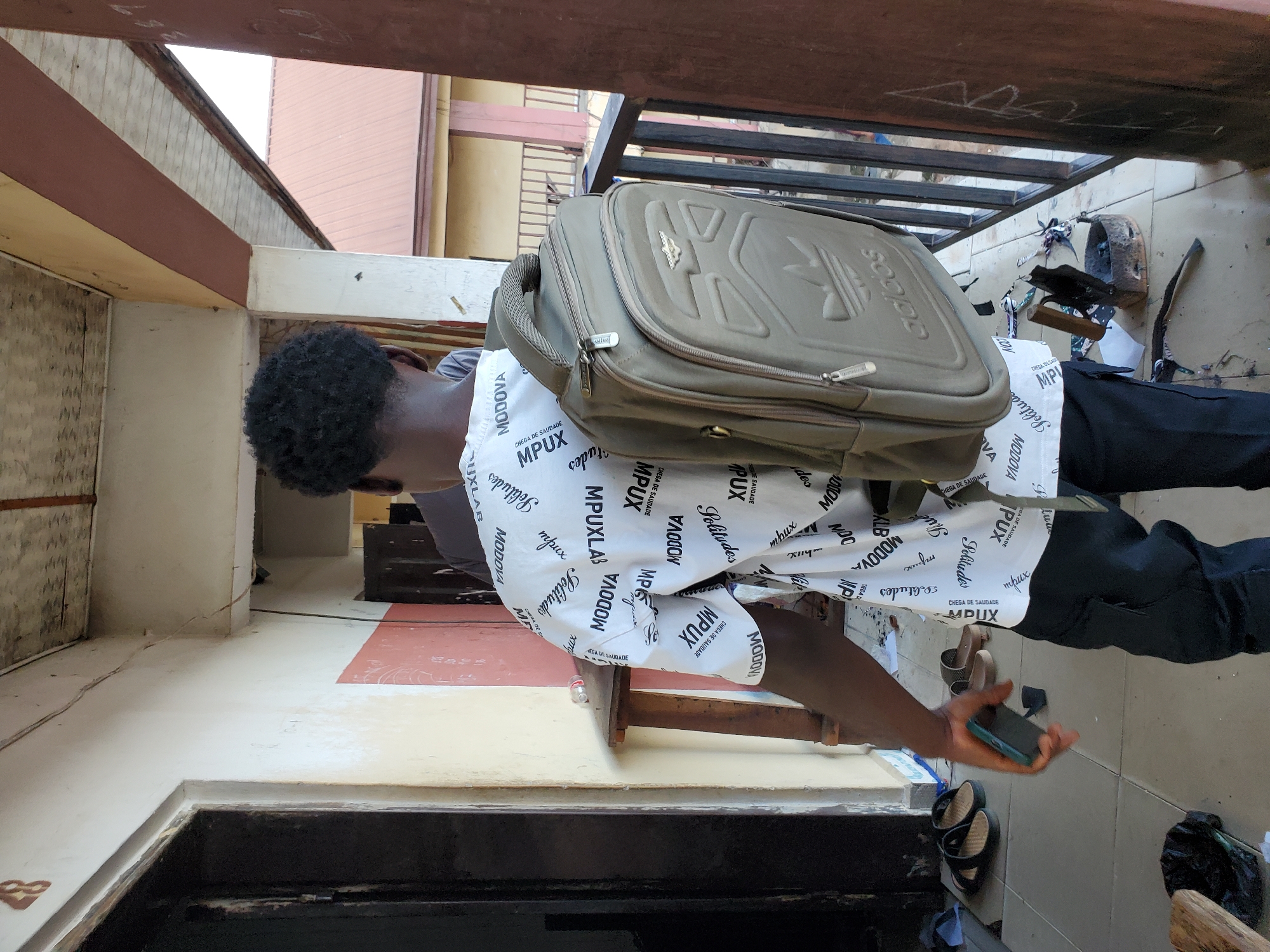 A medium shot captured indoors, though the exact location is ambiguous, shows a person with dark skin and Afro-textured hair wearing a white shirt with black text patterns (MODOVA, MPUX, Solitudes) and black pants, standing near a doorframe or opening. They are carrying a large, grey Adidas backpack. The person is holding a green smartphone in their right hand, seemingly looking at it. The ground and lower walls are tiled, and there are some sandals and a black bag visible near the bottom of the frame. A wooden structure is above and to the right of the person, with miscellaneous items like an old iron and some papers scattered around. The orientation of the photograph has been rotated, so the view appears to be sideways.