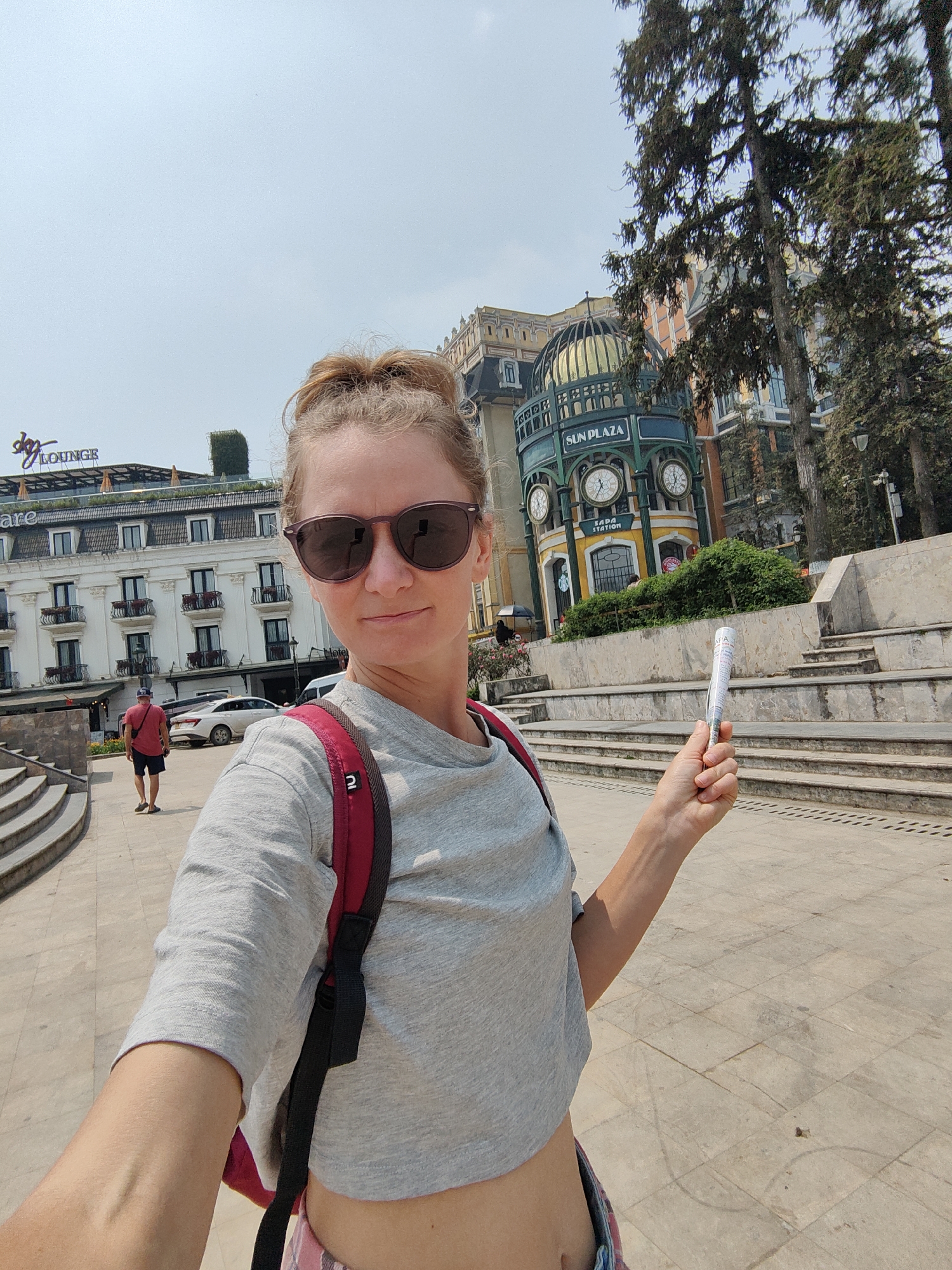 A woman takes a selfie outdoors in Sa Pa, Vietnam, under a clear sky. She is centrally positioned, wearing dark sunglasses, a light grey crop top, and a red backpack, with blonde hair pulled into a bun. Her right hand holds a white cylindrical object, possibly a rolled-up map, with "SAPA" text visible. She has a slight smile. In the background, to the left, is a multi-story white building with balconies and a "dq LOUNGE" sign on its roof. In the center-background stands the Sun Plaza building, characterized by a green and gold domed roof, multiple clocks, and "SUN PLAZA" and "SAPA STATION" signage. Tall evergreen trees frame the right side of the background. A male figure, wearing a red t-shirt and dark shorts, walks away from the camera in the mid-ground, between the woman and the white building. The ground is a light-colored paved surface with ascending concrete steps on both sides.