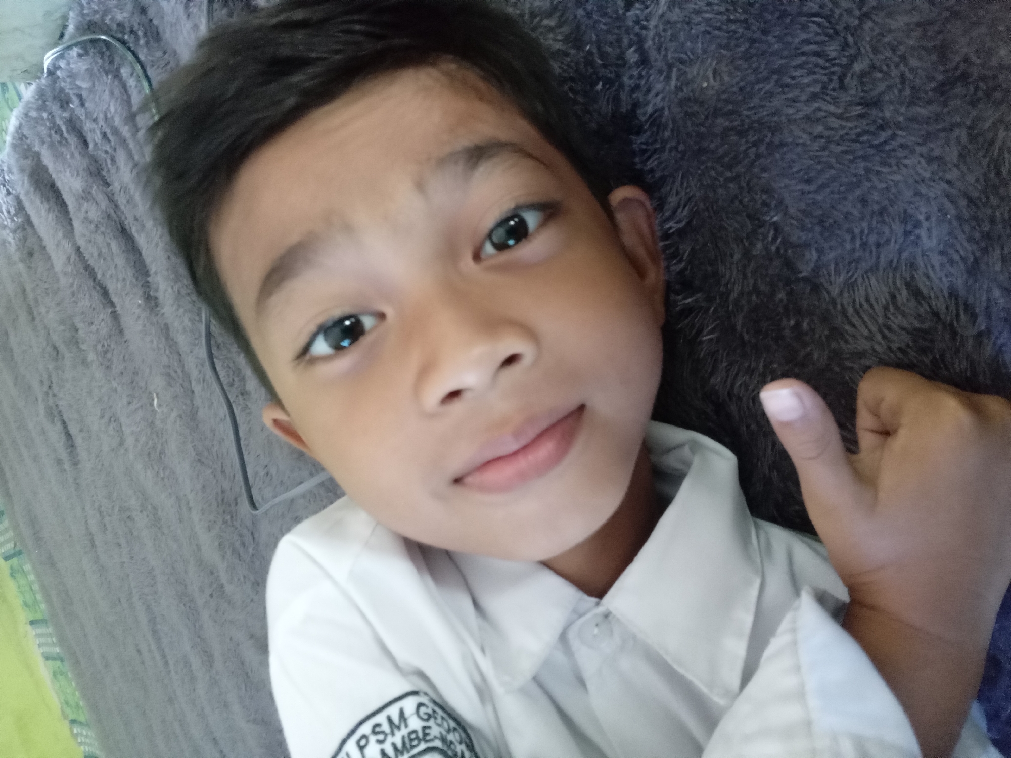 In Kedunggudel, Indonesia, a young boy is pictured lying down on a gray, fuzzy blanket. He is wearing a white collared shirt with a patch on the left side that has some lettering visible. The boy is looking directly at the camera with a slight smile and his left hand is extended forward with his thumb up. The background is a blurry green and gray patterned surface.