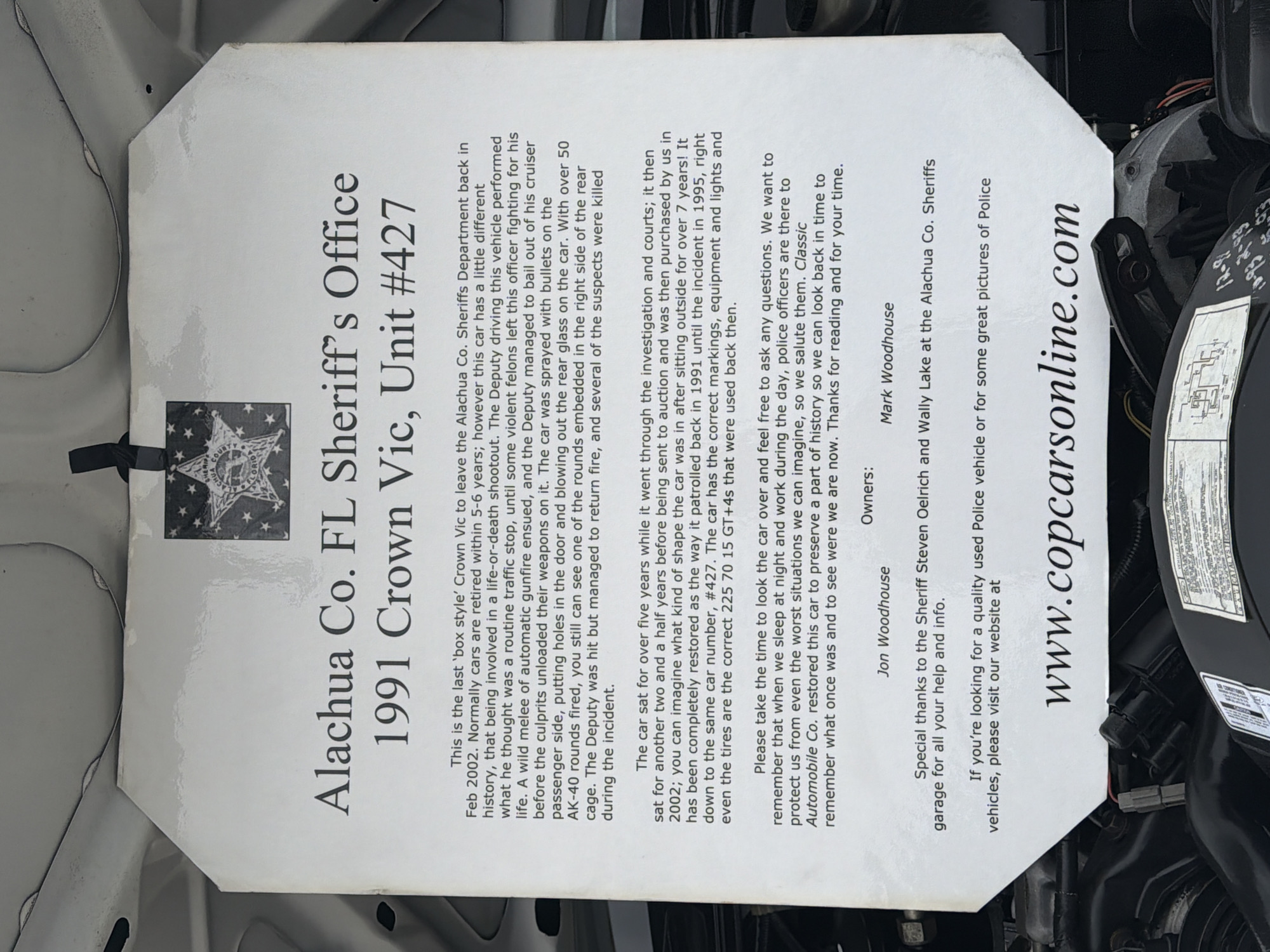 The image shows a sign about a 1991 Crown Victoria, Unit #427, of the Alachua County, Florida Sheriff's Office. The sign details the car's history, including its involvement in a shootout in 1995 and subsequent restoration. The sign includes the names Jon Woodhouse and Mark Woodhouse as owners, and acknowledges Steven Oelrich and Wally Lake at the Alachua County Sheriff's garage. The sign also includes the website address www.copcarsonline.com. The sign is propped up under the hood of a car, with parts of the car's engine visible around it. The location is not specified, but the Sheriff's Office mentioned is in Alachua County, Florida.
