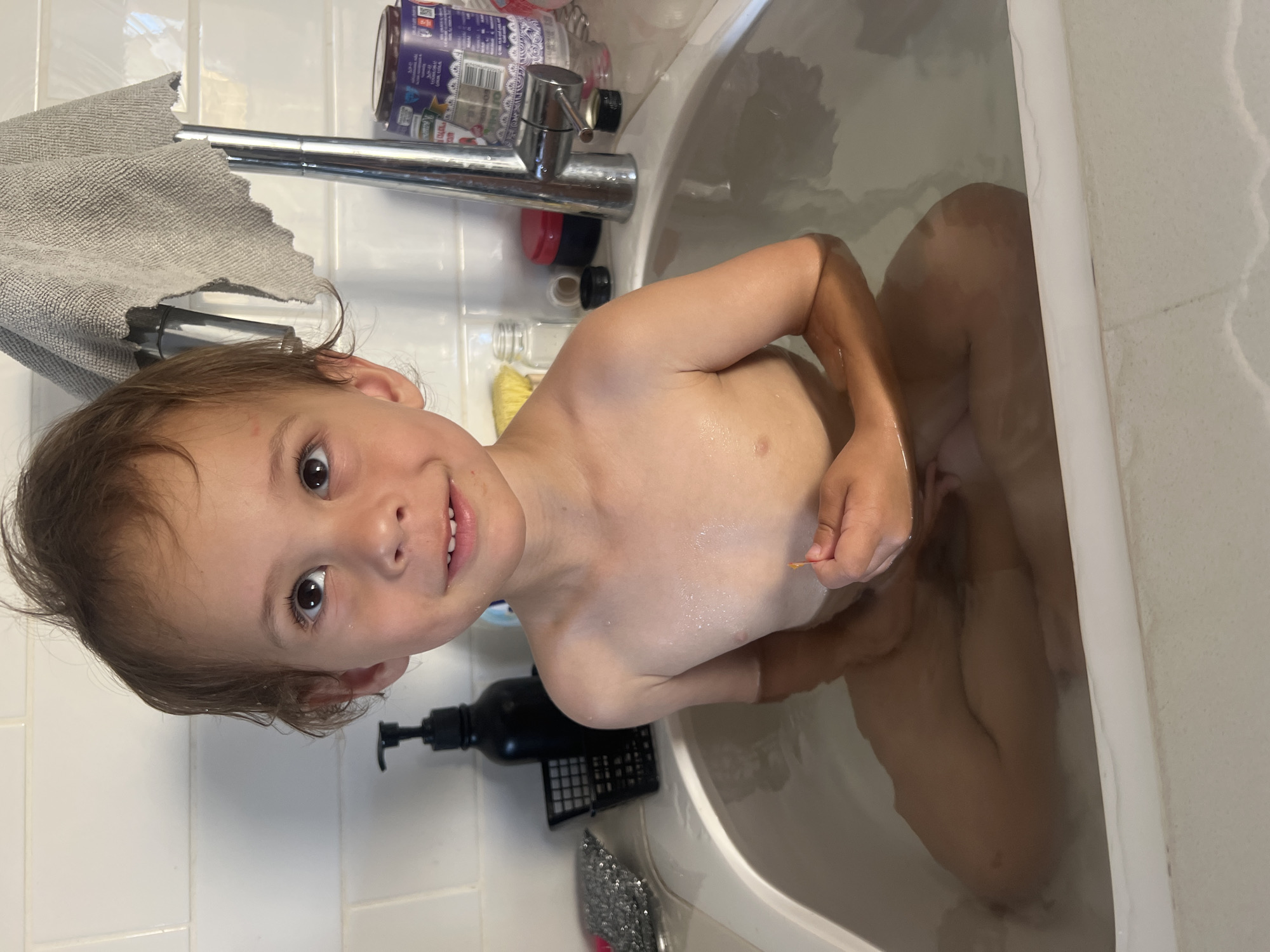 A young child, likely between two and four years old, is the main subject of this photo. They are seated in a white bathtub filled with water, seen from an overhead, slightly angled perspective. The child has light brown hair and dark eyes, looking directly at the camera with a slight smile, revealing some teeth. Their bare torso is visible above the waterline, showing wet skin. In their right hand, they appear to be holding a small, thin, orange or red object.
The bathtub is white, and the surrounding bathroom walls are covered in white rectangular tiles. Above the tub, a chrome faucet with a single handle is visible. To the left of the faucet, a gray, textured towel hangs over what appears to be a shower rail. Various bath products are scattered on the edge of the tub or on the tiled wall behind the faucet, including a clear bottle with a purple label and a black pump bottle. Another small, yellow-bristled brush is also visible near the faucet.
The reflection of the child's body can be seen in the water, indicating the water is clear. The lighting suggests an indoor setting, likely daylight or artificial light. There is no visible text that is clearly legible. The child appears happy and engaged, suggesting a calm and playful bath time scene. Given the location context provided, this scene is taking place in Kiryat Bialik, Israel.