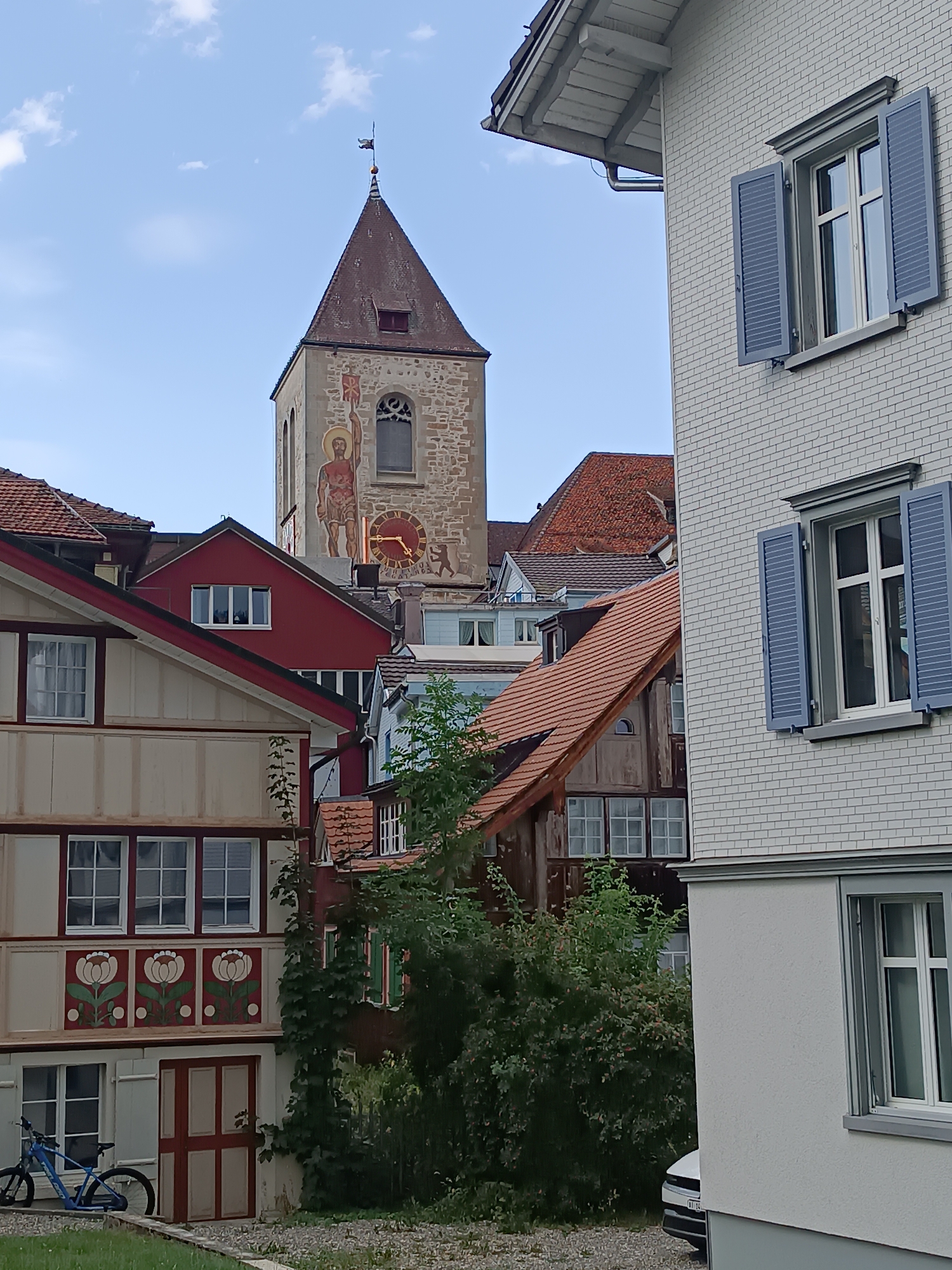 The image depicts a view of the city of Appenzell, Switzerland, focusing on a street scene with historical and modern architecture. Dominating the background is a clock tower of a church, featuring a large clock face and a mural of a religious figure. The tower is made of stone and has a red, pointed roof. In the foreground, there are several buildings with distinct architectural styles. To the right, a modern building with white brick and blue shutters is prominent. To the left, older buildings showcase traditional Appenzell architecture with decorative elements on the facade, including floral motifs and wooden beams. A blue bicycle is parked on the left side of the image, near a red and cream-colored door. Lush green vegetation and trees are scattered throughout the scene, suggesting a well-maintained urban environment. The sky is partly cloudy and blue. The scene appears to be a typical day in the town, with no people actively engaged in any discernible actions.