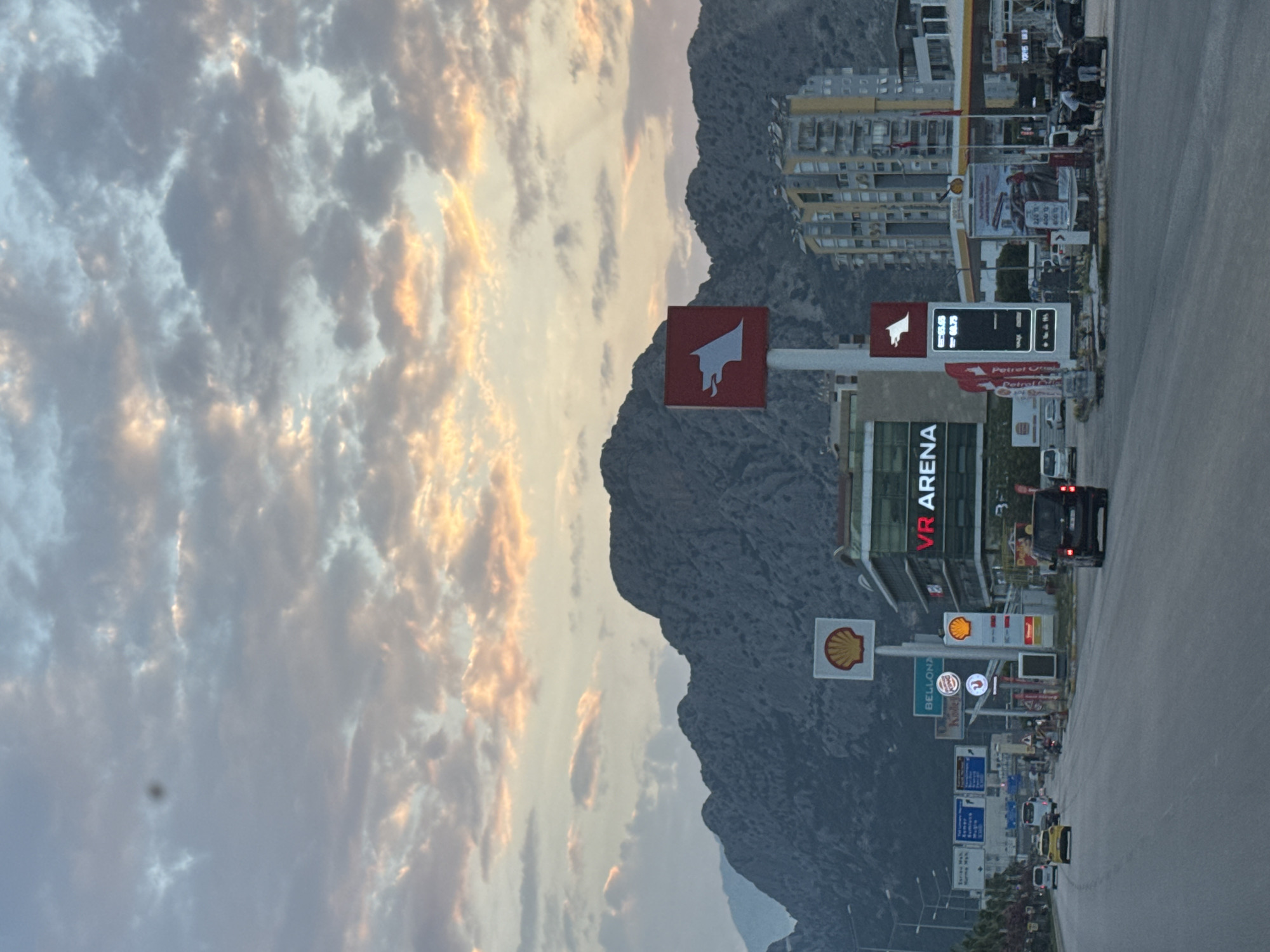 The image captures a vibrant street scene in Konyaaltı, Turkey, during what appears to be either sunrise or sunset, indicated by the warm, golden light illuminating the sky and clouds. The sky, dominating the upper portion of the frame (when upright), is filled with dramatic, textured clouds.
In the background, a majestic and rugged mountain range provides a striking natural backdrop to the urban environment. At the foot of these mountains, various multi-story buildings, some with yellowish facades, are visible.
The foreground features a multi-lane road with several vehicles, including a distinctive yellow car and a dark SUV, suggesting ongoing traffic. Along the roadside, a lively commercial area is depicted with numerous businesses and large signs.
Visible text and logos include multiple "Shell" gas station logos, large red letters spelling "VR ARENA," a blue sign for "BELLONA," and the "Burger King" logo. There are also signs for "Petrol Ofisi," recognizable by its red bull head logo, displaying digital fuel prices such as "28.99," likely indicating Turkish Lira per liter. Other smaller, indistinct Turkish text is present on various directional and promotional signs. The scene portrays a bustling urban setting framed by dramatic natural scenery under a colorful sky.