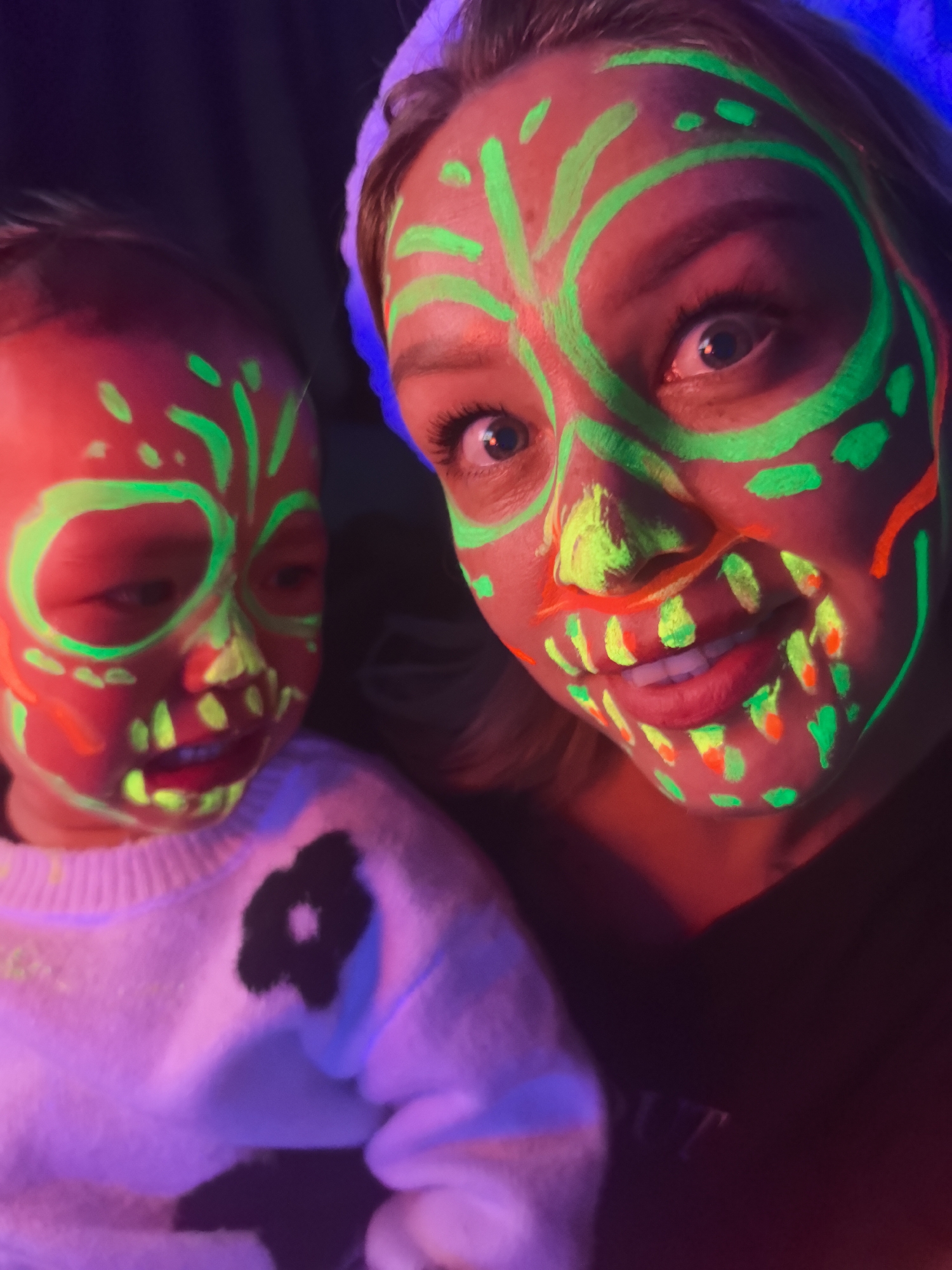 A close-up shot depicts two individuals, presumably a mother and child, under blacklight. Both have fluorescent green and orange face paint resembling a stylized skull. The woman is slightly ahead of the child, with a portion of her black shirt visible. The child wears a white sweater with a black floral design. The background is dark, adding to the effect of the fluorescent paint. "OUT" is faintly visible on the woman's shirt. The image's location is unknown and cannot be deduced from the scene. Noviy, Russia is not discernible from the image.