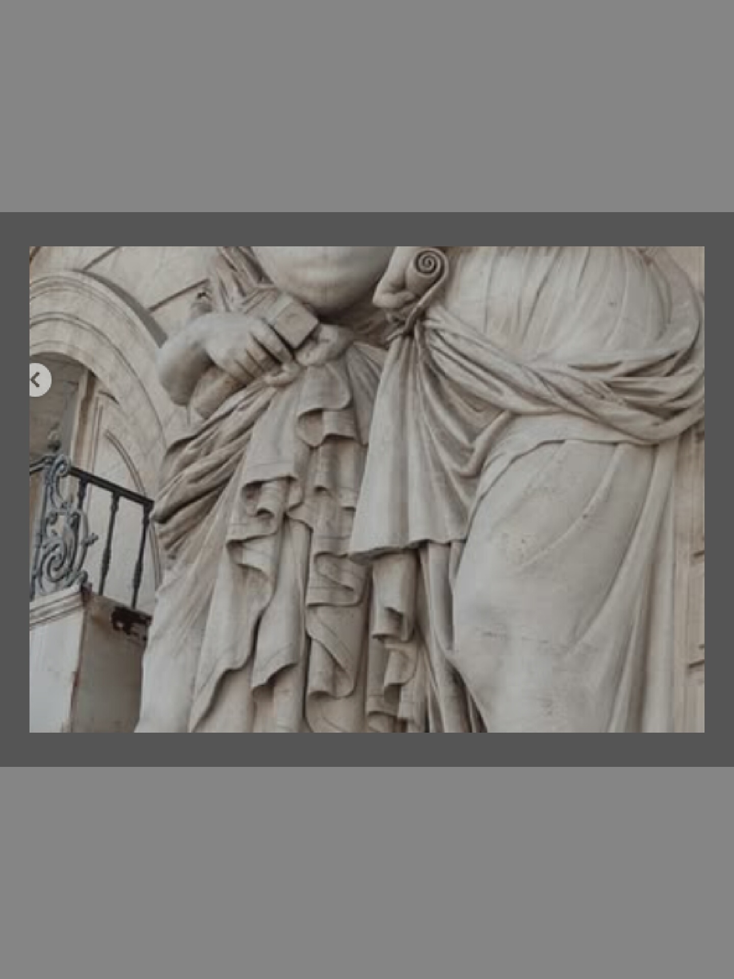 The image shows a close-up view of a stone sculpture of two figures, likely women, draped in flowing robes. The sculpture is rendered in a classical style, with detailed folds and textures in the drapery. One figure's hand is visible, holding a small, rectangular object. The sculpture appears to be an architectural element, possibly part of a building facade, as suggested by the presence of a balustrade and arched stonework in the background. The lighting is natural, highlighting the three-dimensional quality of the carving. The color palette is monochromatic, dominated by shades of grey and white, consistent with stone.