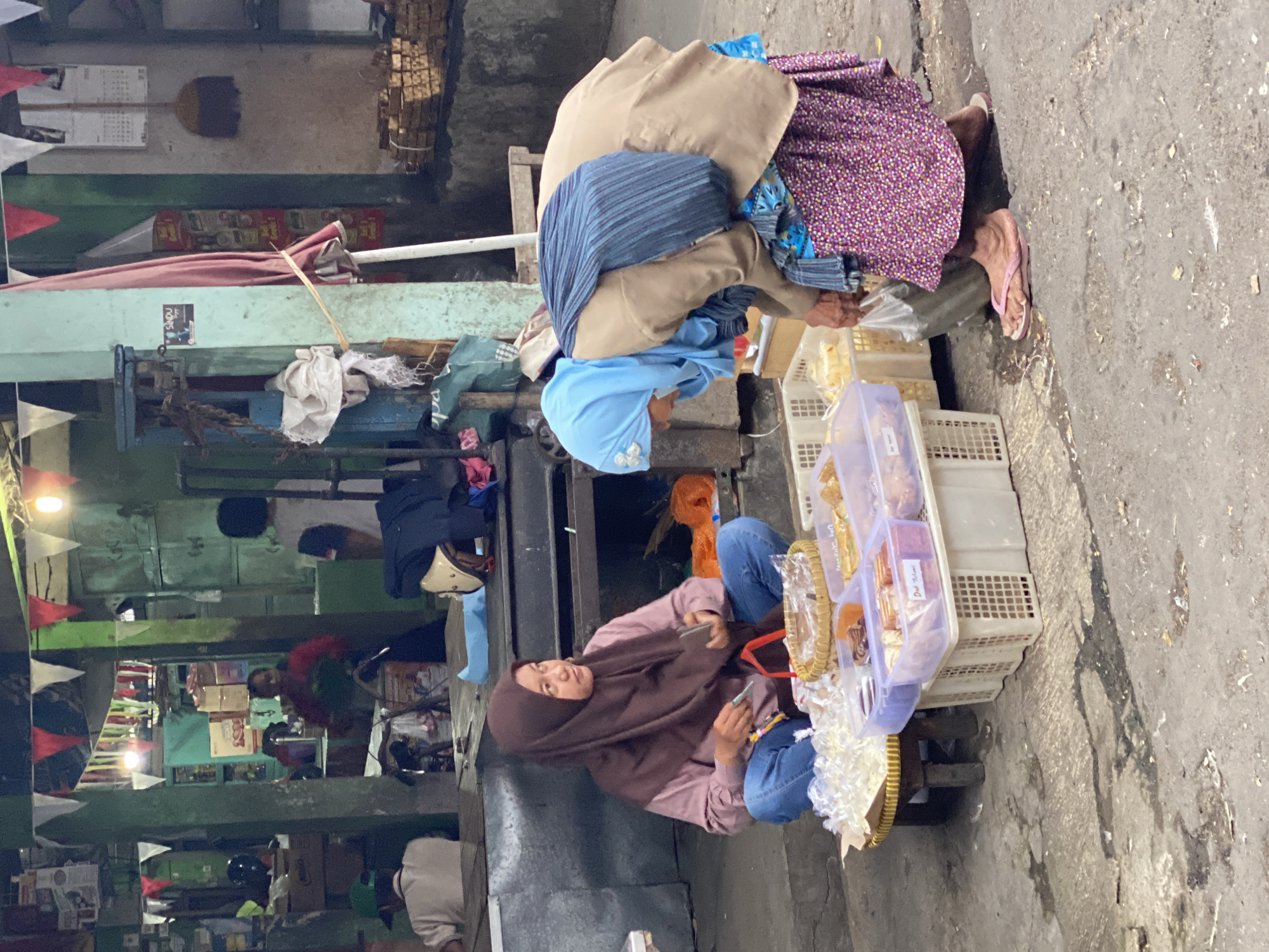 In Ambarketawang, Indonesia, a street market scene unfolds. Two women are the central figures. One woman, wearing a brown hijab and a pink shirt, sits on the ground, cross-legged, facing right. She is holding a small object and appears to be interacting with it. In front of her is a woven mat holding a basket with baked goods and a pile of white items, likely food items, in plastic bags. Beside the mat are several plastic containers filled with various confections.
The second woman, with a light blue hijab, is bent over, facing left and towards the first woman. She is wearing a tan jacket and a patterned purple skirt. She has a striped blue fabric draped over her shoulders and is holding a plastic bag in her right hand. Her left hand is reaching towards the containers of sweets.
The environment suggests an outdoor market setting with stalls and structures in the background. Colorful flags are strung overhead, and there are various items on shelves and counters in the background, including boxes, bags, and what appears to be a broom. The ground is paved with a rough, textured surface. The overall impression is of a bustling, everyday market environment.