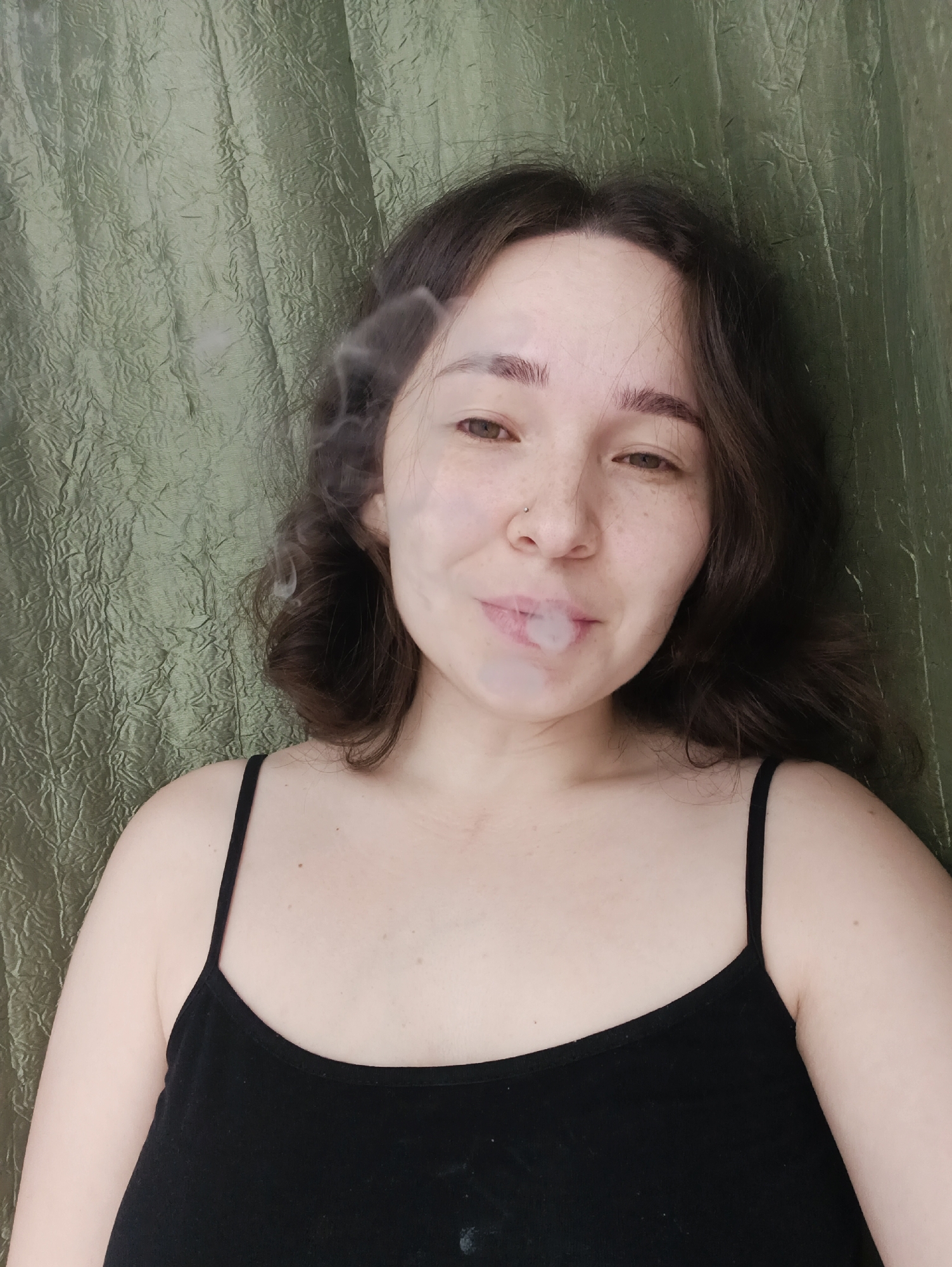 A selfie shot of a young, light-skinned woman exhaling smoke, likely from a cigarette or vape, taken against a textured, green background. She has dark brown hair, brown eyes, and a nose piercing. She is wearing a black tank top. There is no information to deduce the location in the city of Izhevsk, Russia.