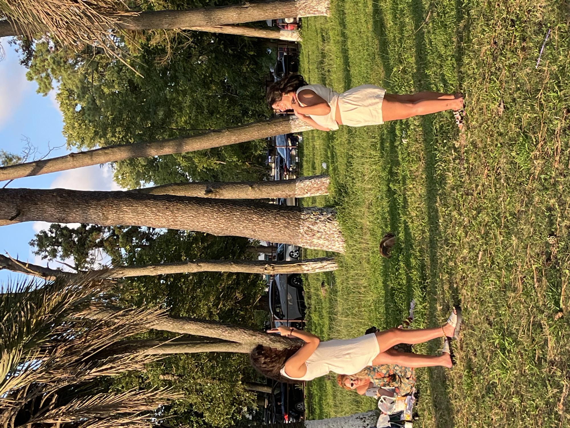 In Ureki, Georgia, two young women are posing for photographs in a grassy park with numerous trees. One woman, on the left, stands facing away from the viewer, wearing a white dress and holding up a phone to take a picture. She has dark, curly hair and is wearing sandals. To her left, a second woman sits on the grass, wearing sunglasses and a patterned top.
Another woman, on the right, stands facing the viewer, wearing a white crop top and white shorts. She is smiling and touching her chest with her hands. She has dark hair and is wearing sandals. In the background, cars are parked, and lush green trees surround the area. The ground is covered in grass with scattered leaves and twigs.