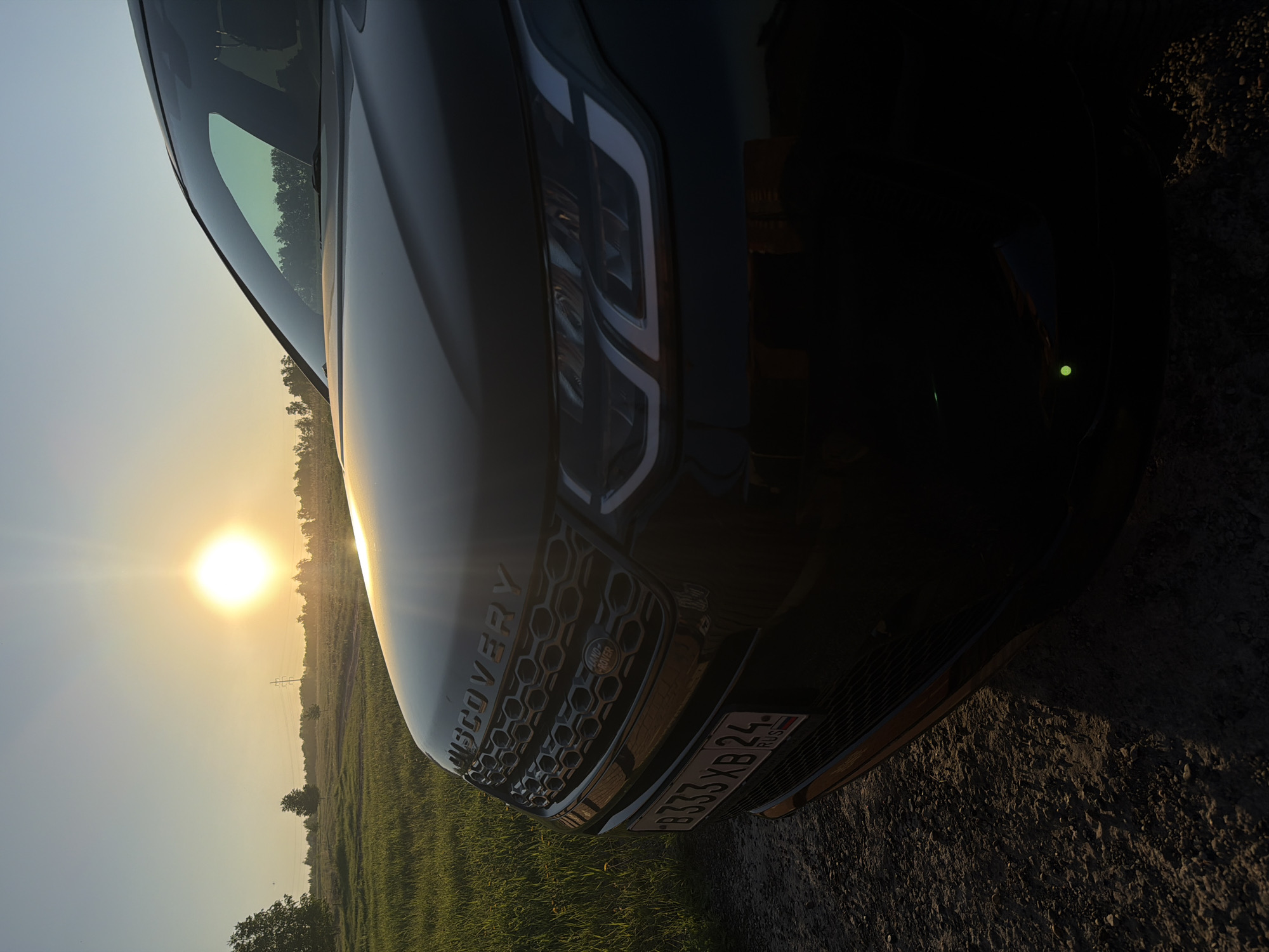 The image captures the front-right quarter of a black Land Rover Discovery SUV, positioned outdoors. The vehicle is parked on what appears to be a dirt or gravel path, with a lush green field stretching out to its left. Trees and utility poles are visible in the distant background of the field.
A brilliant, low-hanging sun dominates the upper-left portion of the frame, casting a warm, golden light across the landscape. The bright, direct sunlight and extended glow suggest either a sunrise or, more likely, a sunset, bathing the scene in soft, golden hues. The sky is clear, transitioning from a light blue overhead to a vibrant yellow closer to the horizon.
Prominently displayed on the car's grille are the words "DISCOVERY" and the "LAND ROVER" emblem. The car's modern headlights are visible, reflecting some of the ambient light. A Russian license plate, reading "B333XB 24 RUS," is clearly attached to the front bumper. The "24" code on the plate signifies Krasnoyarsk Krai, aligning with the provided location context of Krasnoyarsk, Russia. No people are visible in the photograph, which conveys a sense of peaceful solitude amidst a scenic, natural setting.