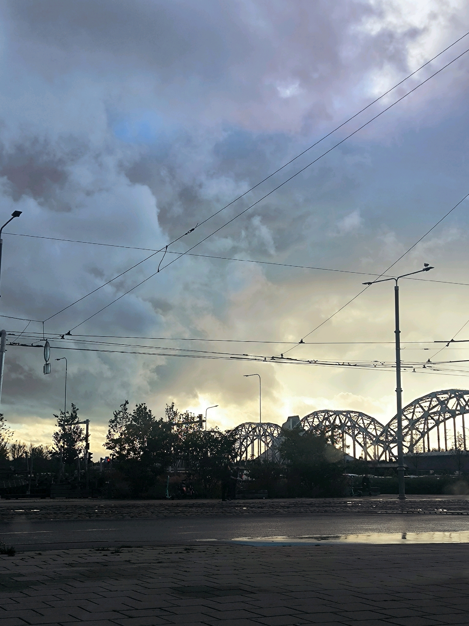 The image captures a street-level view of the Daugava River area in Riga, Latvia, during what appears to be late afternoon or early evening. The sky is dramatic, filled with heavy, dark clouds that are beginning to break up, allowing shafts of golden sunlight to pierce through. These clouds are tinged with grey and blue hues.
Dominating the scene are several overhead wires, likely for trams or trolleybuses, crisscrossing the frame. Two prominent lampposts, typical of urban street lighting, are visible. The foreground is a wet asphalt road, with puddles reflecting the light, suggesting recent rain. The pavement to the left is made of rectangular bricks laid in a pattern.
In the middle ground, a line of trees with dark green foliage forms a silhouette against the sky. Behind the trees, the distinctive arched structure of the Daugava Railway Bridge is clearly visible, its metalwork illuminated by the receding sunlight. The bridge spans across the river, hinting at the broader cityscape beyond.
There are no distinct people clearly visible or engaged in any discernible action, though there are shadows and faint shapes that could indicate distant figures or vehicles. The overall atmosphere is one of a transition, possibly from day to night, or from rain to clearing weather, with a strong emphasis on the atmospheric sky and the iconic bridge. The lighting suggests a muted, slightly cool color palette overall, with the warm sunlight creating striking contrasts.