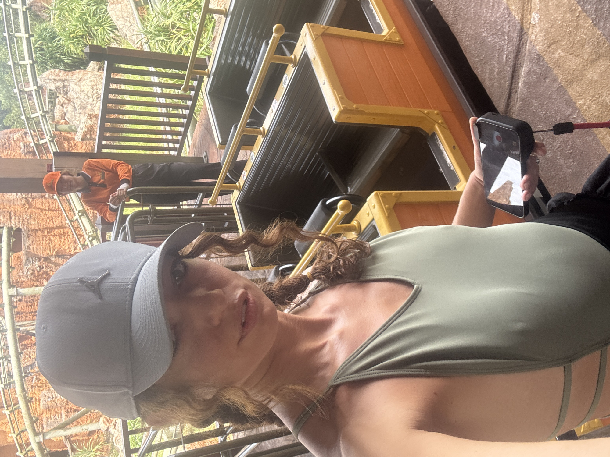 A woman wearing a light gray baseball cap and a green tank top holds a phone in her right hand while seated on a ride. The ride resembles a mine train car with orange and yellow accents and black seats, and another person in an orange uniform stands nearby, possibly operating or overseeing the ride. The background shows a simulated rock and greenery landscape, suggesting an amusement park setting. There's no indication of Đà Nẵng, Vietnam, in the image.