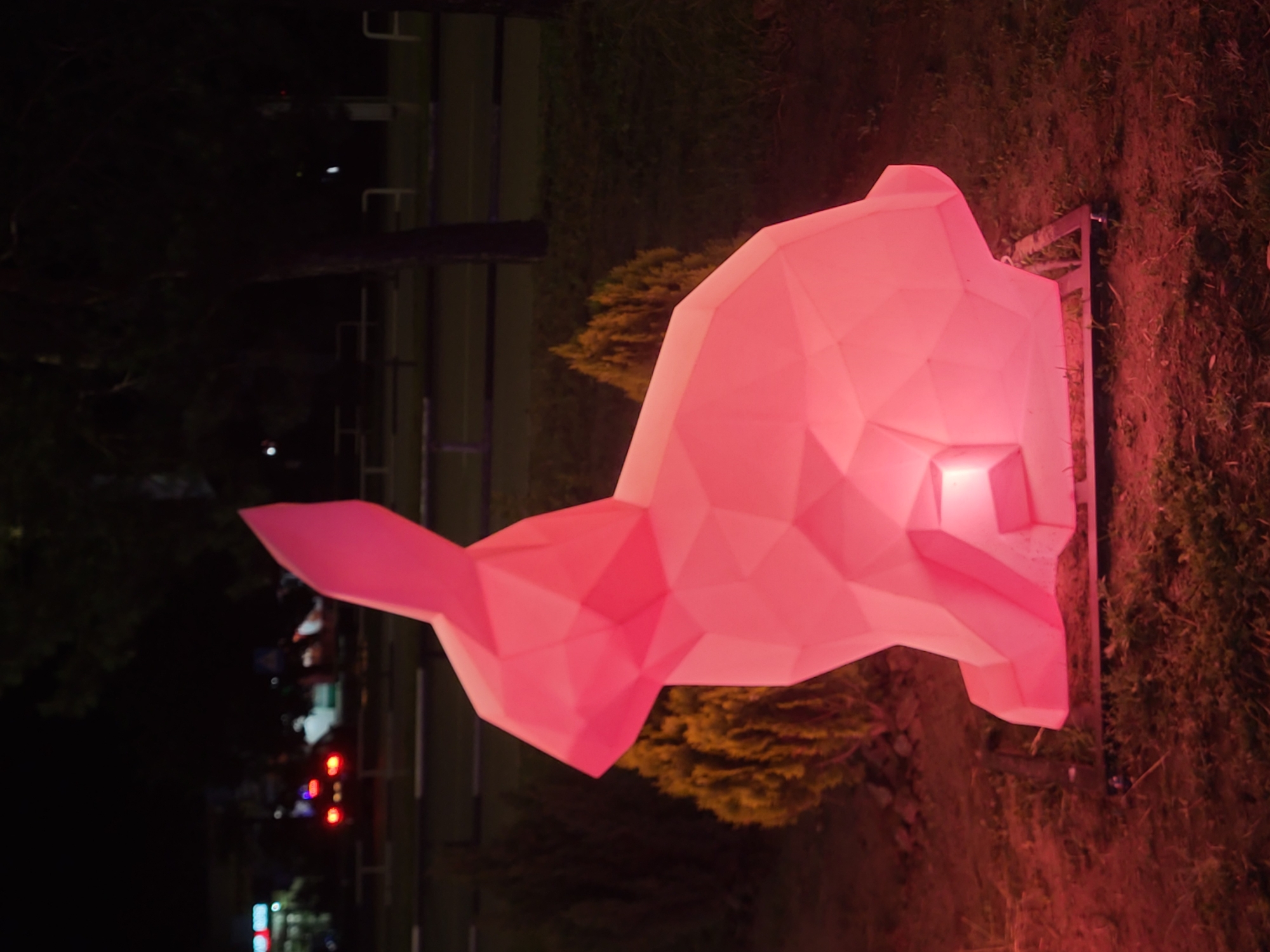 A large, pink, polygonal sculpture of a rabbit is lit from within and positioned on a metal frame in a grassy area at night. The sculpture is the primary subject of the image. In the background, trees and a dimly lit street with cars and buildings are visible. The environment appears to be an outdoor urban or suburban setting. The location is identified as Horishni Plavni, Ukraine.