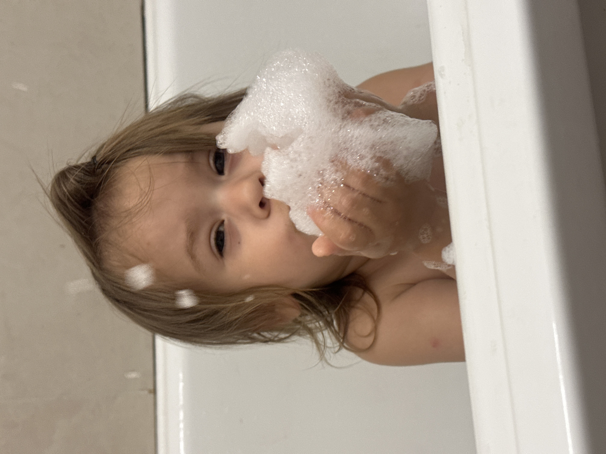 A young child is in a bathtub filled with soapy water and bubbles. The child is holding a handful of bubbles close to their face. The location of this image is unknown.