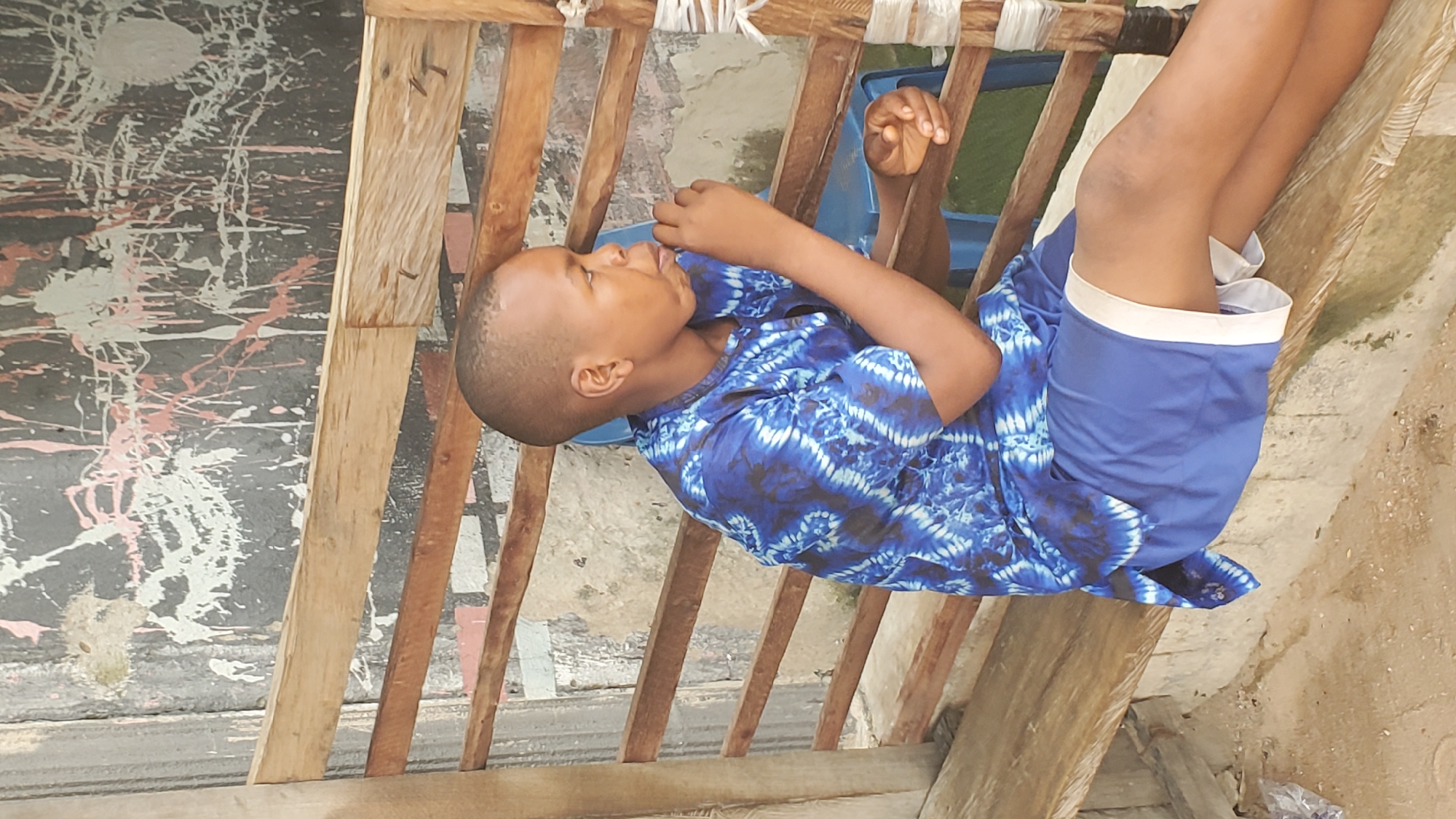A young boy with short hair and dark skin is sitting on a wooden bench in an outdoor setting. He is wearing a blue tie-dye shirt and blue shorts with a white trim. His right hand is near his mouth, and his gaze is directed to his right. He is seated next to a blue plastic chair. The background features a rough, weathered wall with splatters of paint in white and pink. Wooden slats are arranged horizontally behind the boy and the bench. The ground in front of the bench is dirt or concrete. The scene appears to be in Jalingo, Nigeria.