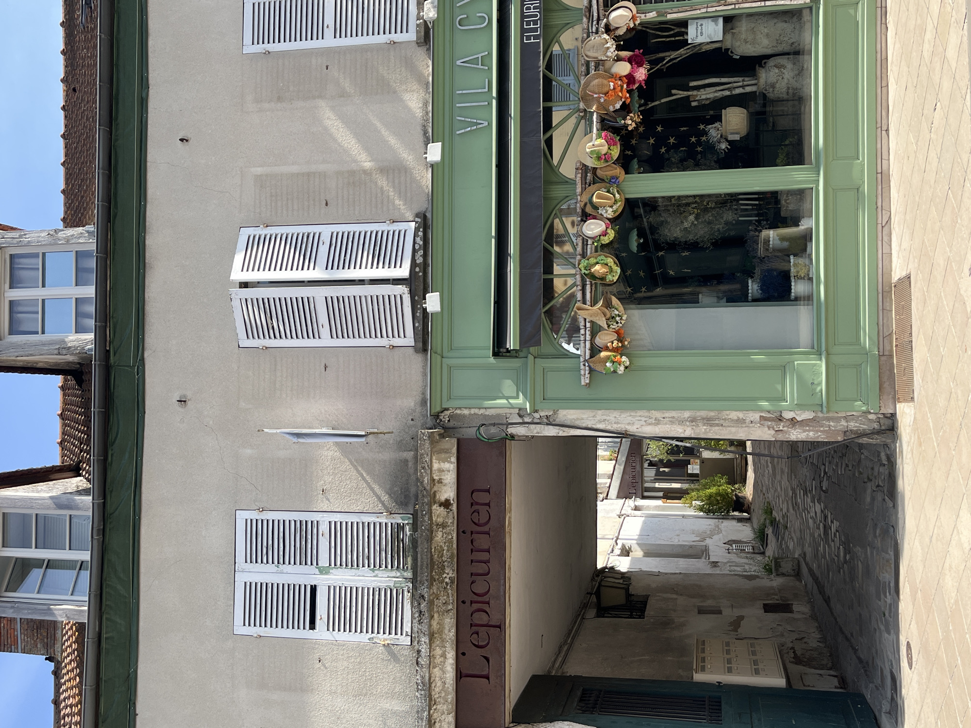 This is a street-level, eye-level, medium shot of a building with two adjacent storefronts in Rambouillet, France. On the left is an archway that leads to a cobblestone alley. Above the archway is a sign that reads "L'Epicurien." On the right is a green storefront with large windows displaying flowers and plants. A sign above the windows reads "VILA CY" and below that "FLEURISTE". Several decorative hats with flowers are hung above the storefront windows. The building has light-colored stucco walls and white louvered shutters on the windows. The sky is clear and blue.
