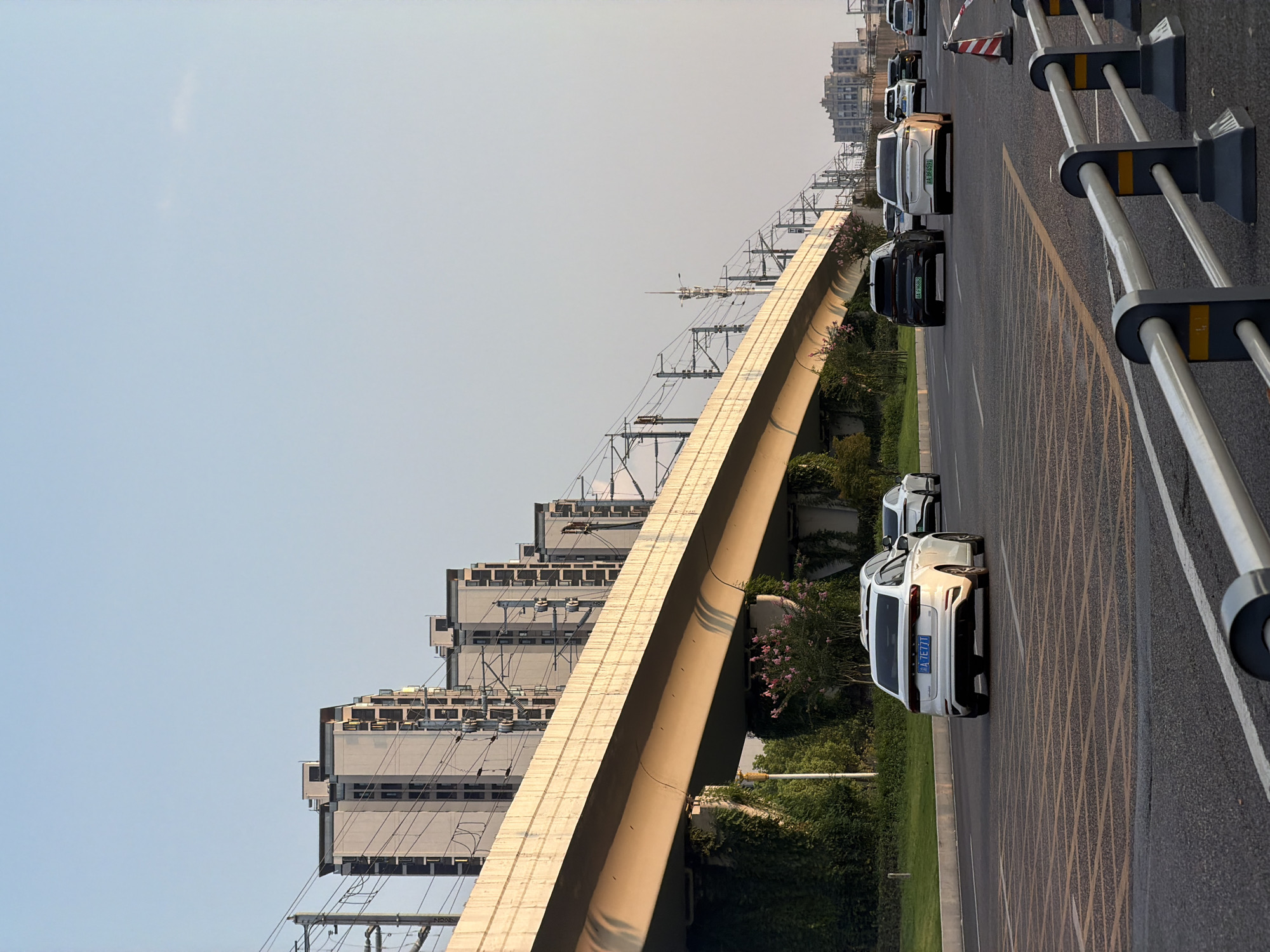In Jiangjin, China, a three-lane asphalt road runs horizontally across the foreground. The road is marked with yellow diamond-shaped patterns and white lines. Guardrails with white and dark gray posts line the right side of the road. Several cars are traveling on the road. In the center of the frame, a white sedan with a blue license plate (A 7E771) is driving away from the camera. To its left, a black SUV with a green license plate (A F8682) is also moving away. Further down the road, more cars are visible.
Above the road, an elevated concrete structure, likely a bridge or overpass for a railway or highway, extends from left to right. The underside of the structure is supported by concrete pillars covered in green ivy. Delicate pink flowers bloom on bushes beneath the overpass. Power lines and train tracks are visible along the top of the overpass, stretching into the distance.
In the background, a row of modern, multi-story apartment buildings with a light beige facade and dark windows is visible on the left side of the frame. On the right, a cluster of taller buildings suggests a more urbanized area. The sky is a clear, pale blue with a faint haze, indicating it is likely late afternoon or early evening. The overall scene depicts a typical urban landscape with infrastructure and residential areas.