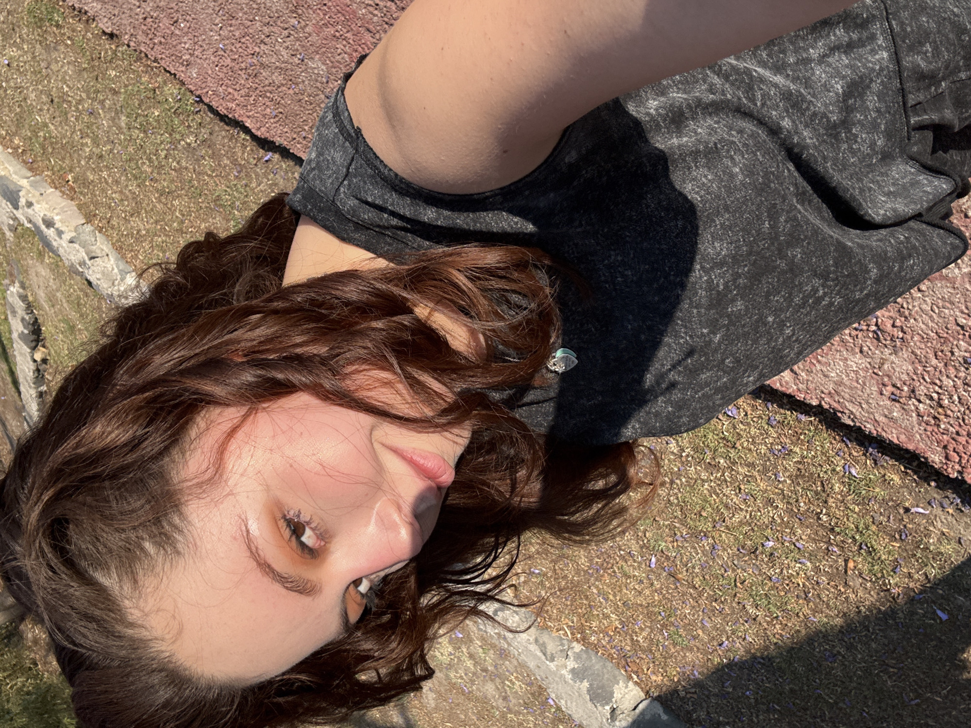 A young woman is lying on her back in an outdoor setting in Santa Clara Ocoyucan, Mexico. Her head rests near a weathered stone structure, while the rest of her body lies on a surface partially composed of red paving stones and partially of dry grass with scattered purple flower petals. She wears a dark gray top. The image is taken from an overhead perspective, casting a shadow across the lower portion.