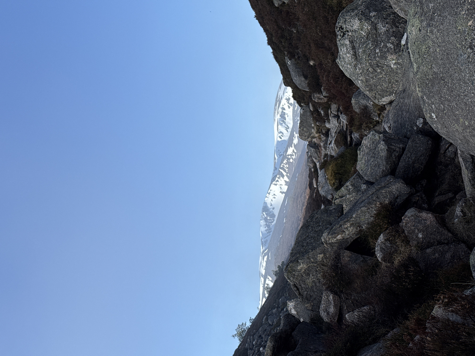 The image captures a mountainous landscape under a clear blue sky. Dominating the right side are large, gray rocks interspersed with dark soil and vegetation. Beyond the rocks, a snow-capped mountain range is visible. The location and city are unknown.