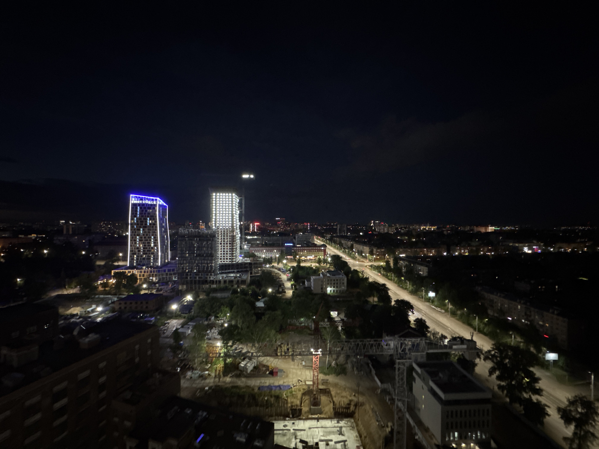 An aerial, night-time view of a construction site and urban area in Izhevsk, Russia. In the foreground, a large building under construction is visible with exposed concrete foundations and scaffolding. A red tower crane is erected on site, with parts of another crane visible to the right. Several smaller buildings and trees are scattered throughout the scene. In the mid-ground, a wide road with streetlights runs horizontally across the image, with cars visible on it. On the left side of the road, a cluster of modern high-rise buildings are illuminated, one with a blue neon outline. Further in the distance, the city extends with more buildings and lights, under a dark, cloudy sky.