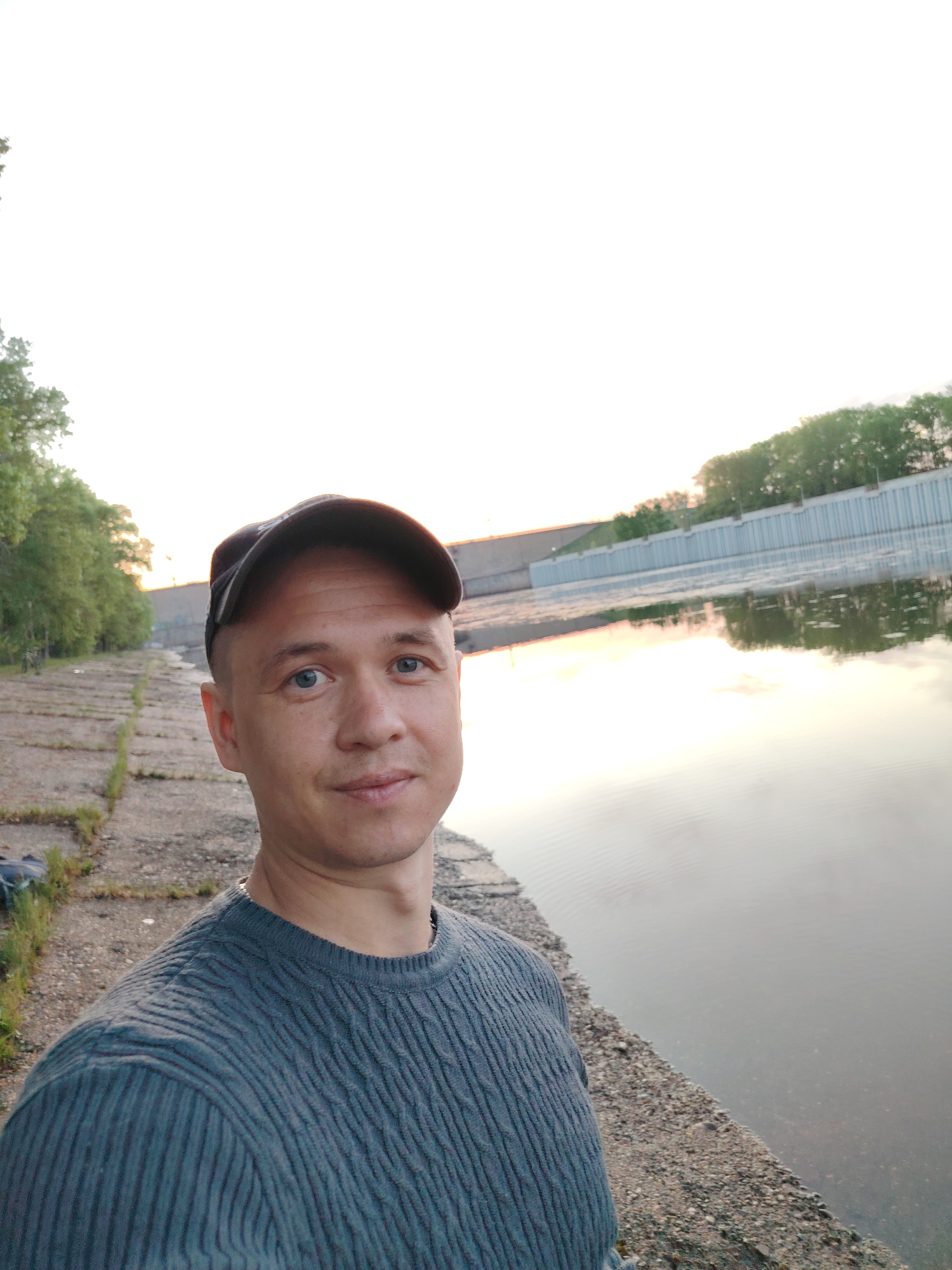 A man is taking a selfie. He is wearing a dark-colored baseball cap and a blue ribbed long-sleeved shirt. He has blue eyes.
He is standing on a concrete and gravel edge next to a body of water, which appears to be a river or lake. Behind him, across the water, is a concrete structure with trees lining the top edge. To his left is a concrete walkway with vegetation growing in the cracks. The sky is light and suggests it might be dusk or dawn.
The location is Chaikovsky, Russia.