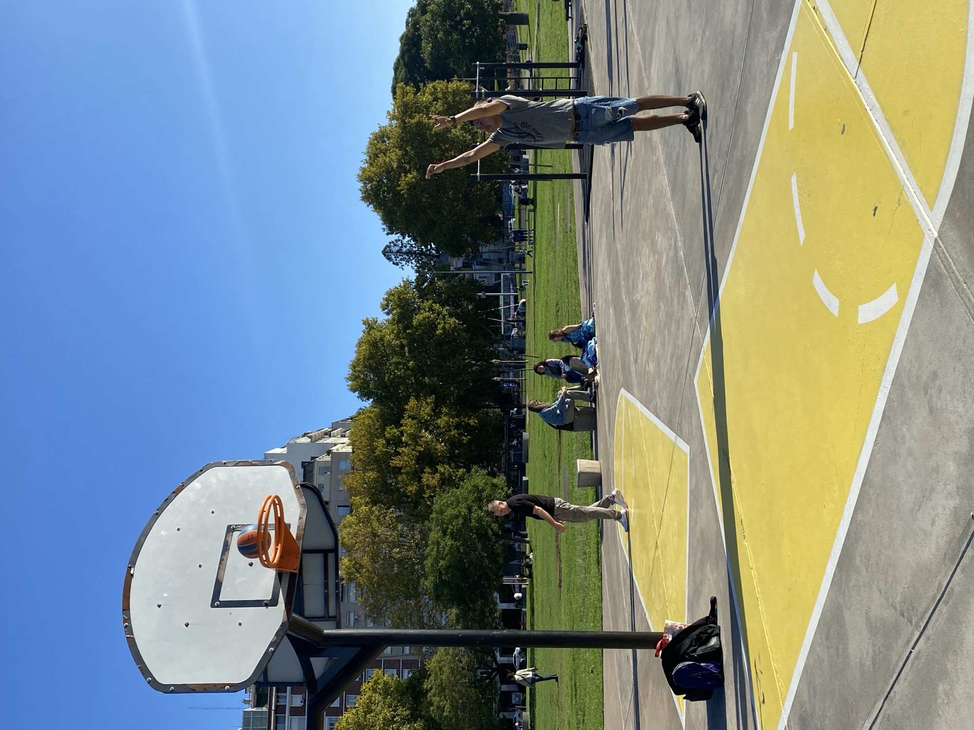 The image captures a scene on a sunny day at a basketball court in Buenos Aires, Argentina. The court features a yellow and grey painted design. A man wearing a grey t-shirt and denim shorts is on the court with his arms outstretched. Another man in a black shirt is standing near the basketball hoop. A group of people is seated in the background. The basketball hoop is visible with an orange rim and backboard, and a ball lodged in the net. Trees and buildings surround the court. A black backpack lies near the base of the hoop. The sky is clear and blue.