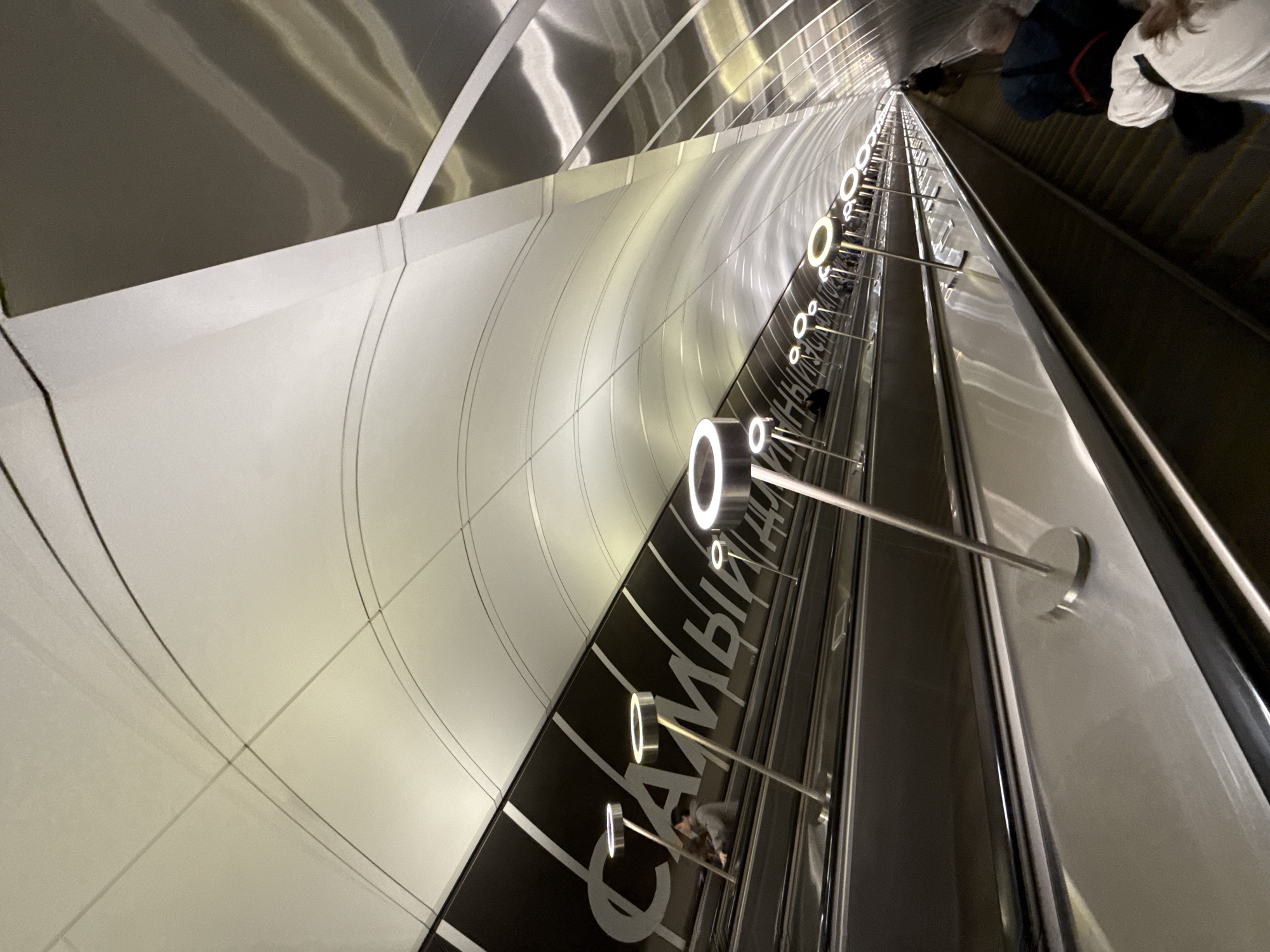 This image captures a modern and impressively deep escalator tunnel within the Moscow Metro, viewed from a high, tilted perspective looking downwards. The long escalator descends into the depths of the station, with numerous steps and moving handrails visible.
The tunnel walls are constructed from a combination of materials, featuring reflective metallic panels on the upper sections that gleam under the light, and lighter, matte-white curved panels on the lower portions. Subtle lines and patterns are integrated into these panels, adding to the contemporary design.
Illumination is provided by a series of circular, glowing white LED lights, mounted on slender silver poles extending from the side wall alongside the escalator. These lights cast reflections on the polished surfaces, enhancing the sense of depth and modern aesthetic.
Several people are visible riding the escalator. In the upper right foreground, two individuals are partially seen from behind; one wears a white top, and the other has a dark blue jacket with a backpack featuring a red stripe. Other passengers are discernible further down the escalator, appearing as smaller figures.
A notable detail is the large white Cyrillic text displayed on a black strip running along the side of the escalator. The visible portion reads "САМЫЙ ДЛИННЫЙ" (SAMYY DLINNYY), which translates to "THE LONGEST." This signage refers to the fact that the Moscow Metro is home to some of the longest escalators in Europe, specifically at stations like Park Pobedy.
The scene conveys the functional yet grand scale of a modern urban transport system, characterized by clean lines, bright lighting, and continuous movement. The indoor setting precludes specific weather or time-of-day details beyond artificial illumination.