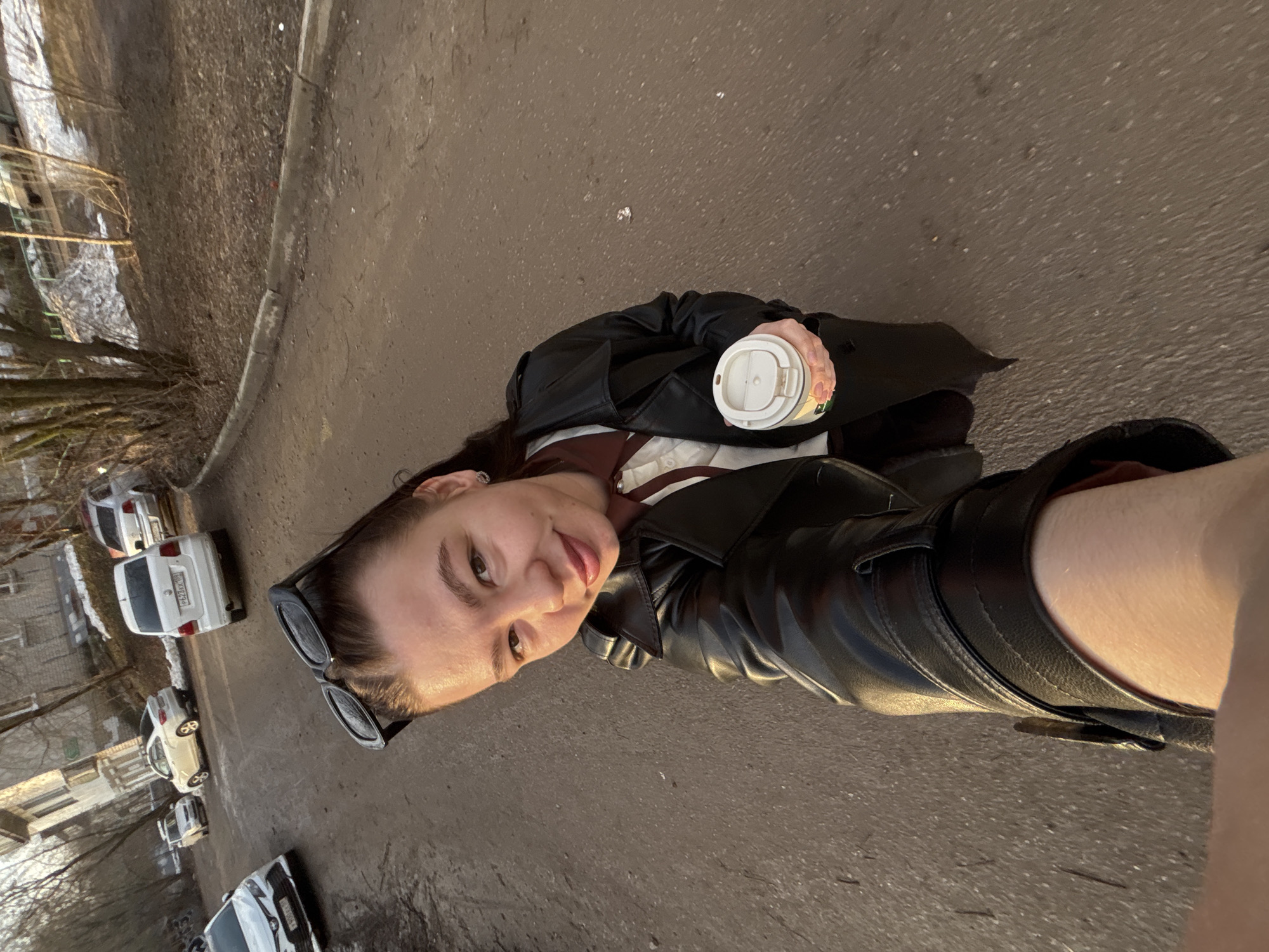A young woman is taking a selfie while standing on a paved sidewalk near a street. She is holding a white coffee cup with a lid in her left hand. She is wearing a black leather jacket and sunglasses resting on her head. The background includes a building, parked cars, and trees. The image was taken in Perm, Russia.
