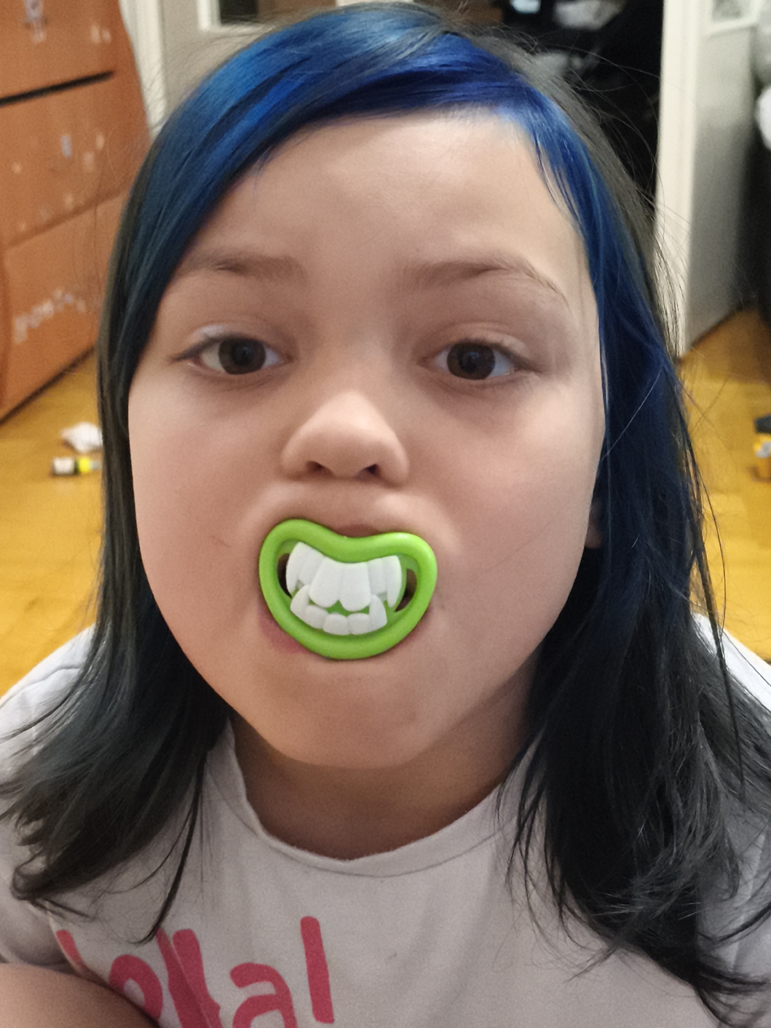 The image shows a close-up of a young person's face, with bright blue-dyed hair. They are wearing a pacifier with fake, oversized white teeth. The background is out of focus, but appears to be an indoor setting with a wooden floor and some furniture. There are no discernible landmarks or details that would indicate the location in the city of Chaikovsky, Russia. The image focuses on the subject and their unusual pacifier, creating a somewhat humorous or playful impression.