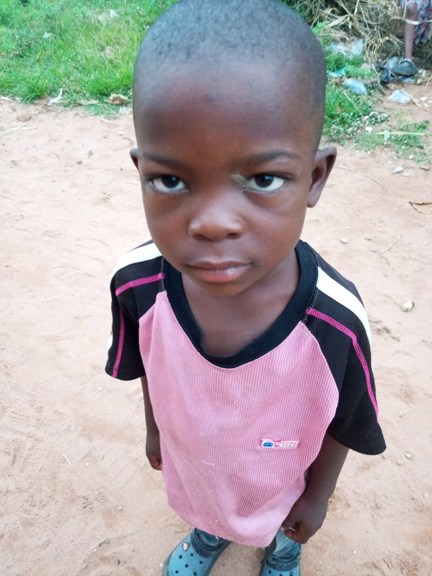 A young Black child, likely a boy, stands on sandy ground in an outdoor environment. The child has a shaved head and is looking directly at the camera with a neutral expression. They are wearing a pink and black t-shirt with white and pink accents on the shoulders and sleeves, and blue Crocs. The background consists of dry grass and some debris, suggesting a rural or unpaved area. The location is identified as Tempure, Nigeria.
