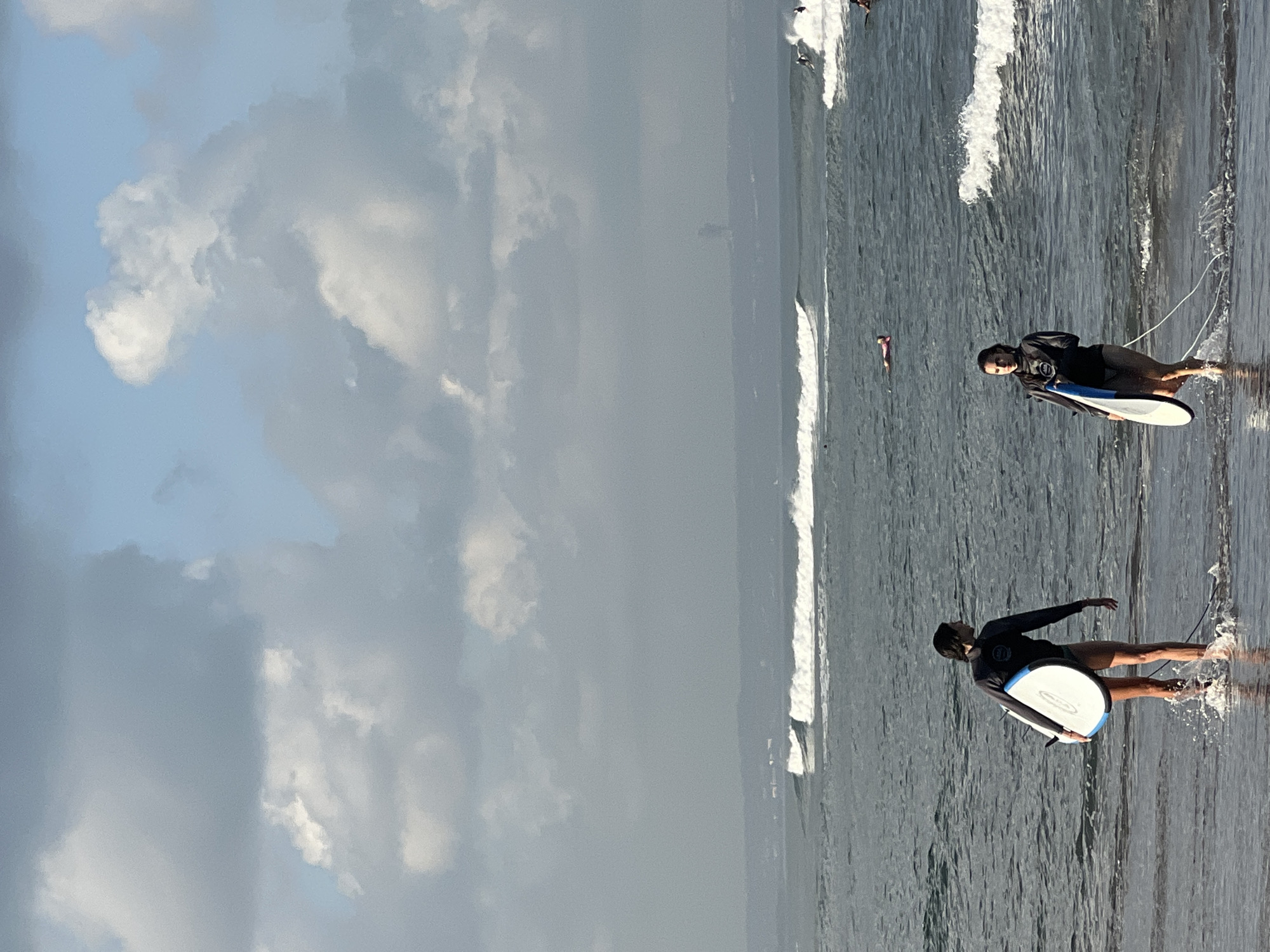 Two surfers, a woman and a man, walk on the wet sand at the edge of the ocean in North Kuta, Indonesia. The man, in the foreground on the left, wears a black wetsuit top and dark swim trunks, carrying a white surfboard with blue trim. The woman, on the right, also wears a black wetsuit top and dark swim trunks, carrying a white surfboard with blue trim. Both are walking away from the camera towards the water. The ocean is calm with gentle waves breaking further out. Several other surfers are visible in the water, some riding waves and others paddling. The sky is partly cloudy with a mix of grey and white clouds against a pale blue sky. In the distance, a hazy shoreline with some structures is visible.