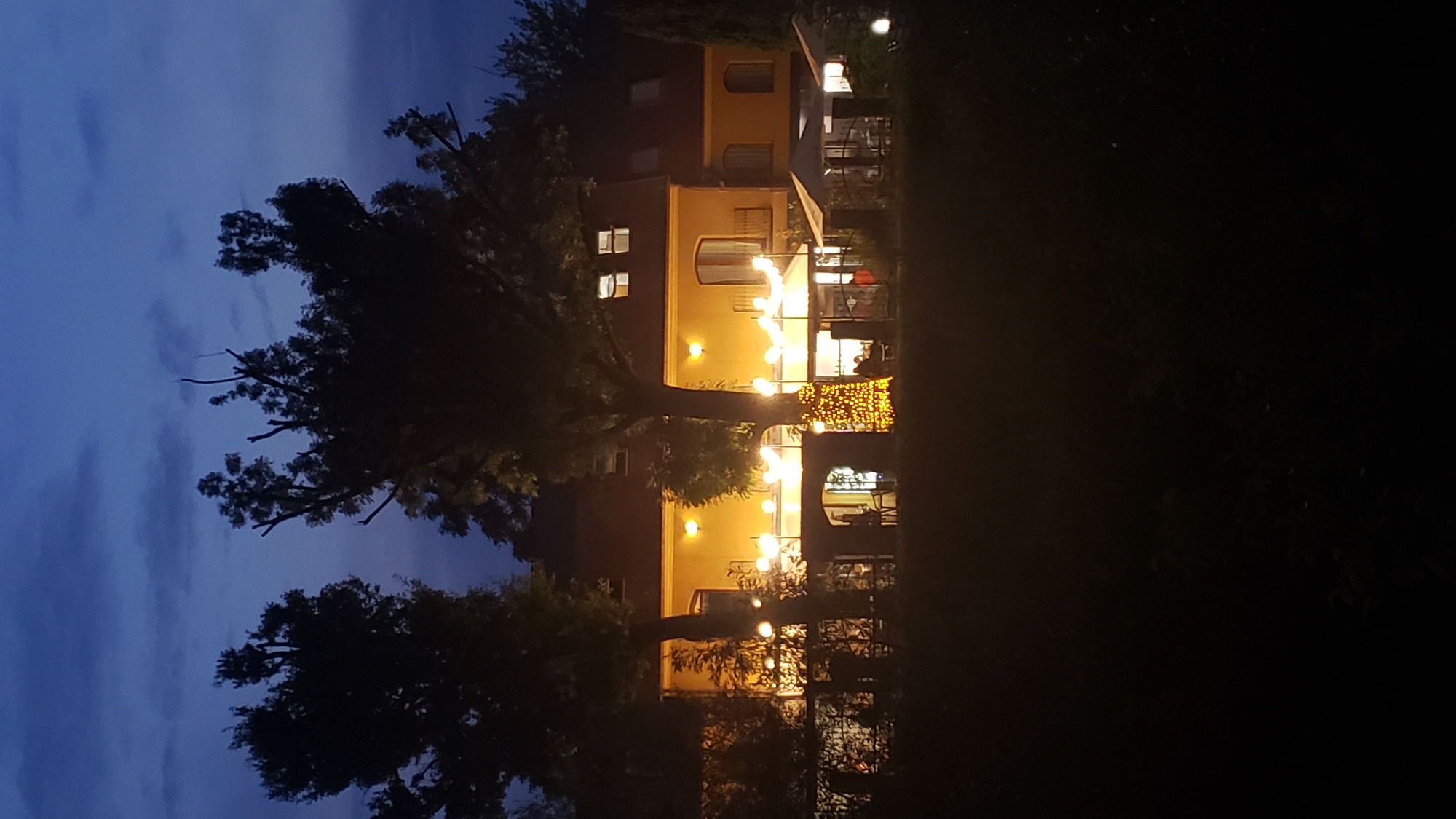 The image depicts a building at night, illuminated by numerous yellow lights. The building appears to be a restaurant or a similar establishment, with outdoor seating areas covered by umbrellas. String lights are draped across the front of the building, and a tree is decorated with festive lights. The sky is dark blue, with a few clouds visible. The scene is set in a dimly lit, natural environment, with dark trees and foliage in the foreground and background. The location is the restaurant "Zámok" in Prešov, Slovakia.