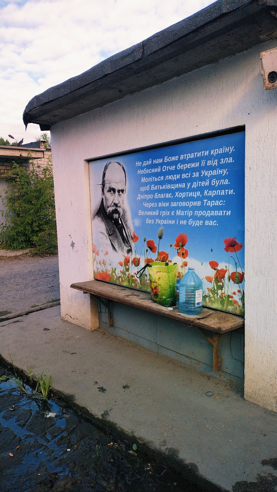 The image captures a close-up of a building facade with a large mural depicting a portrait of a man with a beard and mustache, set against a background of red poppies. Text in Ukrainian is overlaid on the mural. To the left of the mural, a patch of green foliage and a section of a brick wall are visible. A weathered wooden bench is mounted on the wall below the mural. On the bench, there is a green reusable shopping bag with a floral pattern and two plastic water bottles, one large and one smaller. The foreground shows a wet, paved area with some standing water and debris. The overall scene suggests an outdoor, possibly urban, environment. The mural appears to be a patriotic or religious message, likely related to Ukrainian heritage and well-being.