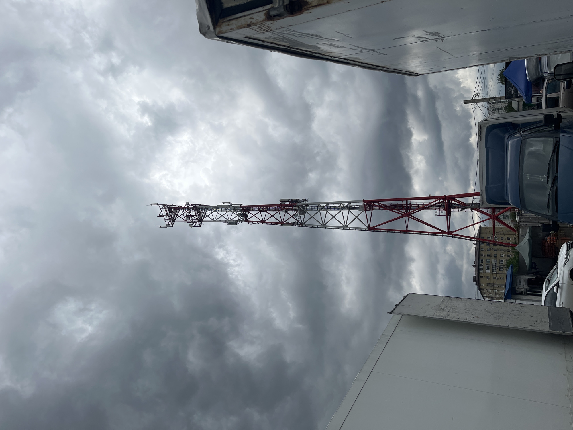 The image captures a view looking upwards between two large vehicles, likely trucks, towards a cell tower against a cloudy, overcast sky. The tower is painted with alternating red and white sections. In the background, a multi-story apartment building is partially visible. The location is likely Krasnodar, Russia, but this identification is solely based on contextual clues. There is also a car parked under blue awning and a glimpse of a building sign "Marazin". The scene appears to be an industrial or commercial area, judging by the presence of the trucks. The photo seems to have been taken from ground level, looking up.
