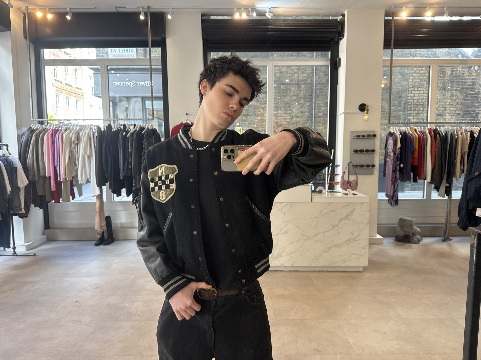 A young man takes a mirror selfie inside what appears to be a clothing store or boutique during the daytime. He has dark, messy hair and a relatively neutral expression, with his head tilted slightly. He is wearing a black varsity jacket with leather sleeves, a black t-shirt, dark jeans, and a brown belt. A patch on the left chest of his jacket displays letters that appear to be "NVCU" or similar, and the number "8". He holds a modern smartphone, likely an iPhone with multiple rear cameras, in his right hand to capture the image.
The background reveals a retail space with light-colored tiled flooring, white walls, and a white marble-patterned counter. Clothing racks are filled with a variety of garments including jackets, shirts, and dresses in colors such as pinks, purples, browns, blacks, and whites. Other objects include sunglasses displayed on a shelf, a small purple handbag on a counter, and a pair of black boots on the floor near a clothing rack. A grey, furry plush toy is visible on the floor in the deeper background.
Large windows behind the clothing racks look out onto an urban street scene with brick buildings. Reflected text in the window, visible in reverse, includes "STREET IW" and "Oliver Spencer", a known menswear brand, along with other partial text that helps identify the location as London, United Kingdom. The store is illuminated by natural light from the windows and overhead track lighting.