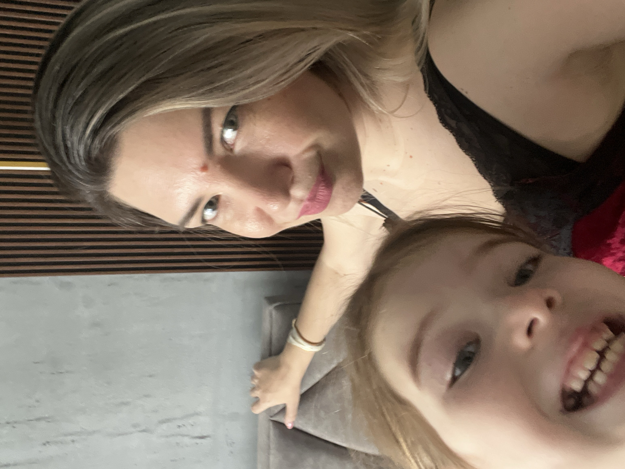 A medium shot selfie shows a woman and a young child. The woman, in the foreground and slightly to the right, has blonde hair and is wearing black lace clothing. She has a red bindi on her forehead and is smiling. The child, in the foreground and to the left, has blonde hair and is smiling widely, showing teeth. The background includes a gray, textured wall and a vertical wooden slatted wall. The woman's left arm is resting on a grey upholstered headboard. The image appears to have been taken indoors, likely in a bedroom. There is no contextual information to place this image in Sochi, Russia.
